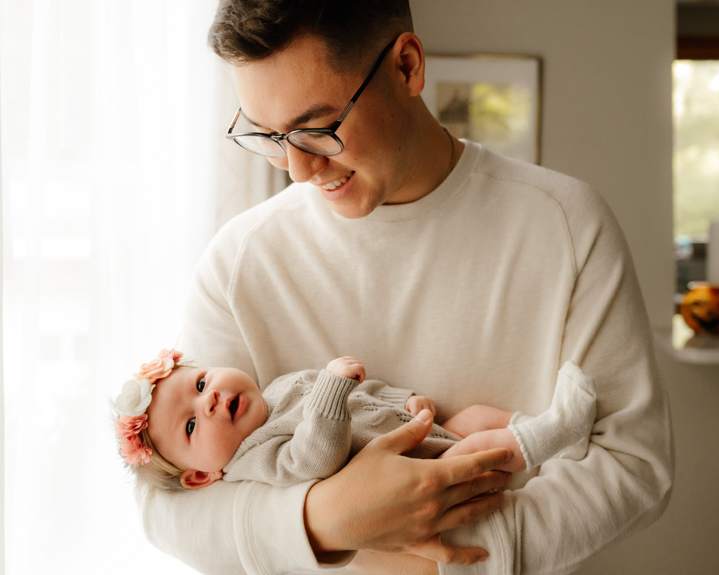 a new father cradling his baby girl in his arms and smiling down at her as she looks directly at the camera during a newborn photography session