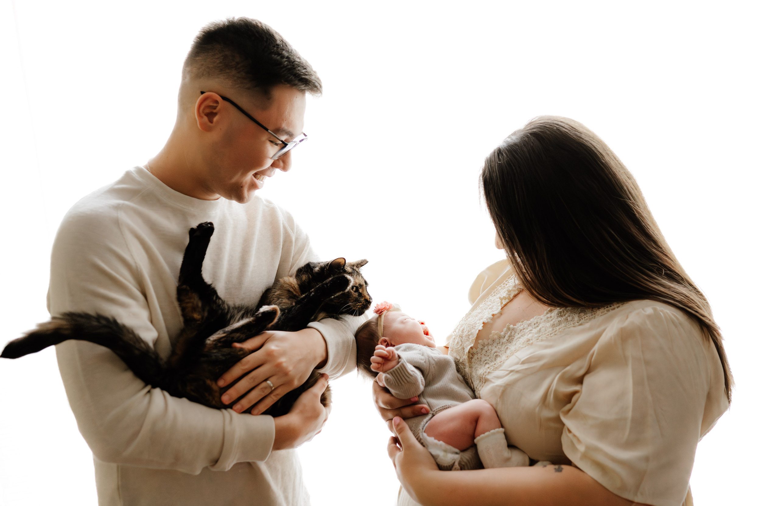 a backlit photo of new parents holding their baby girl and their pet cat in their arms and smiling as the cat sniffs the baby's head during a Glenside newborn photoshoot