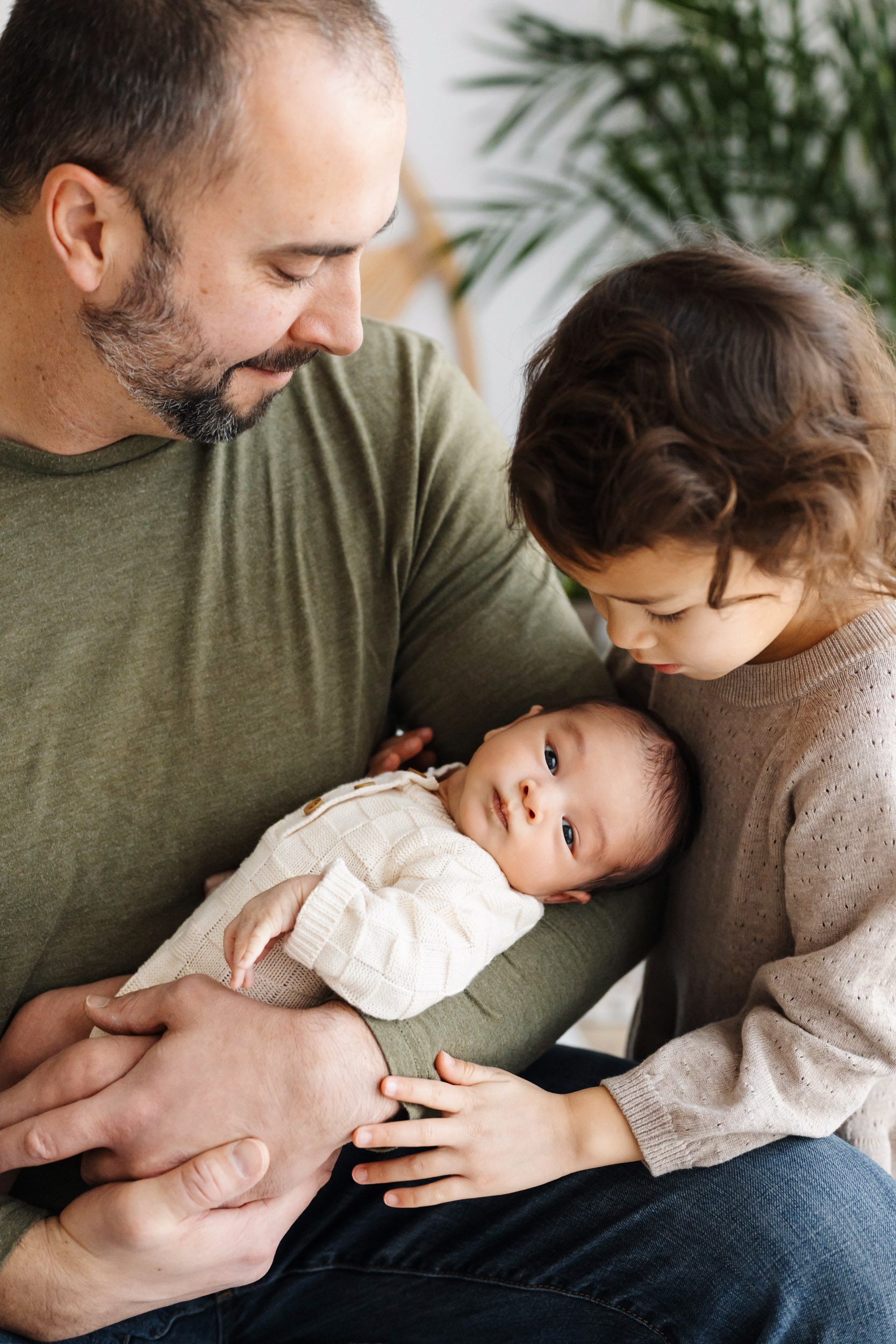 a baby boy cradled in his dad's arms as his big sister stands next to dad and smiles down at her brother during a family centered newborn photoshoot