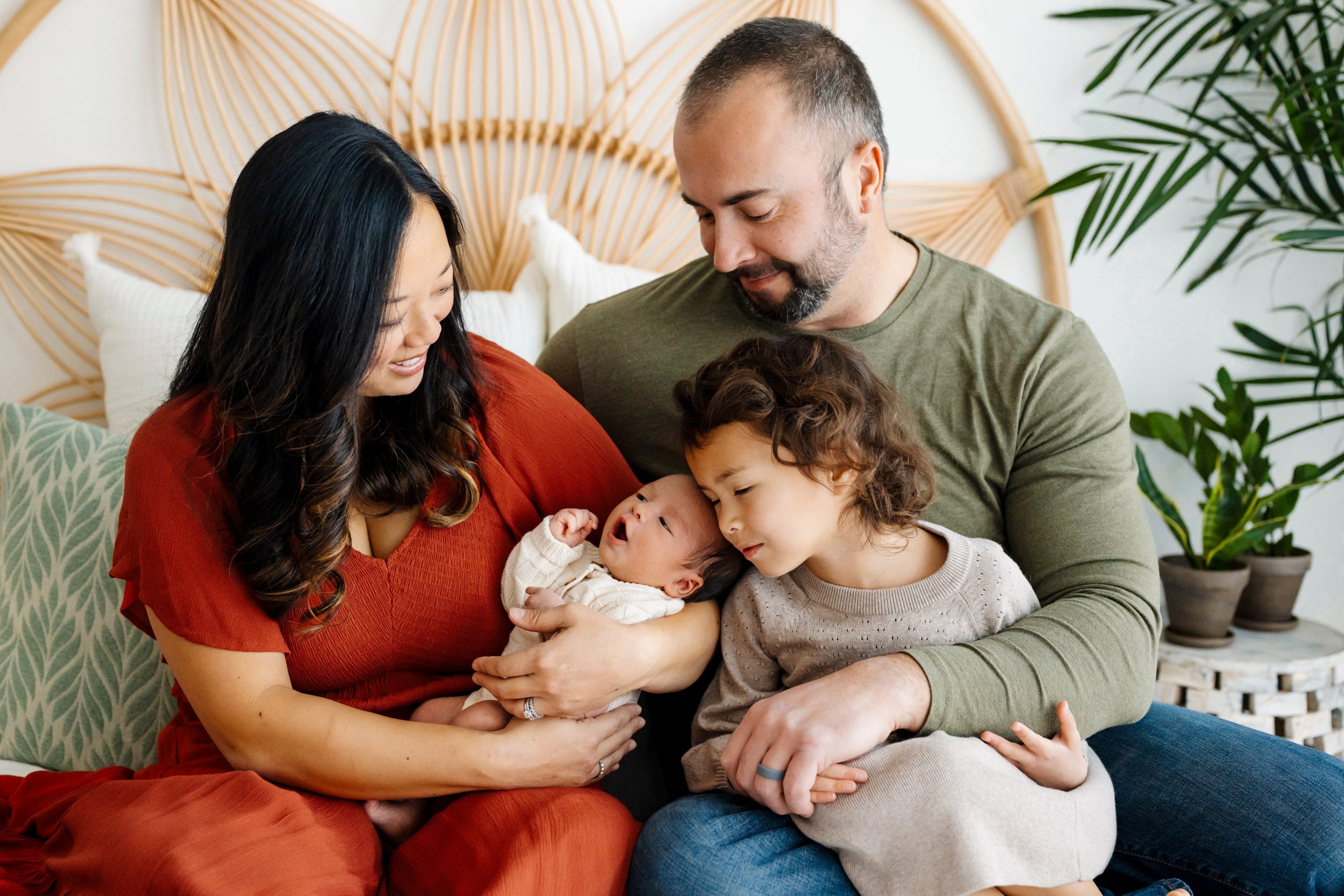 a family of four sitting on a bed and smiling down at their new baby boy cradled in mom's arms while big sister rests her head against the baby during a family centered newborn photo session
