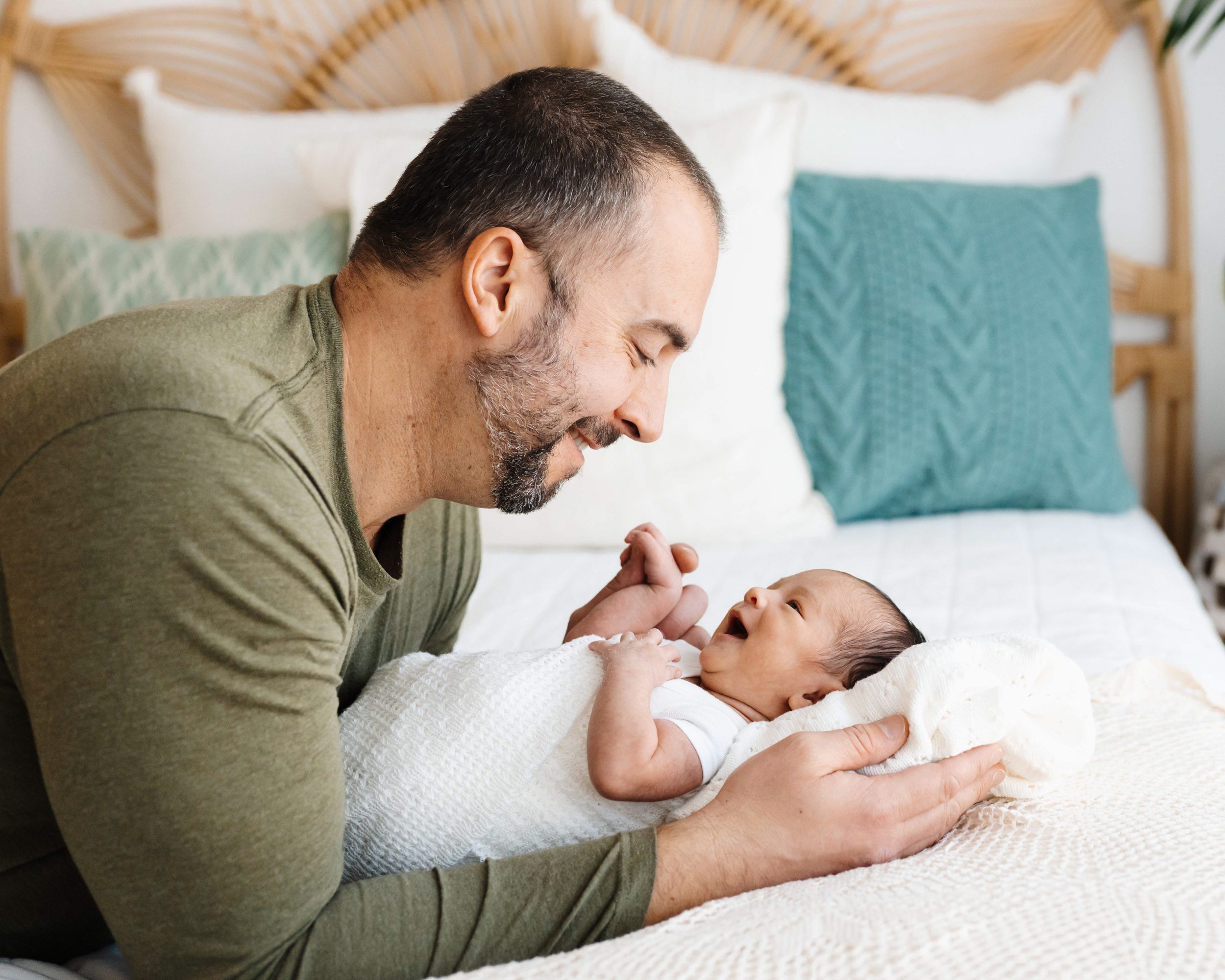 a dad laying on a bed with his baby boy as the baby smiles up at him during a family centered newborn photoshoot