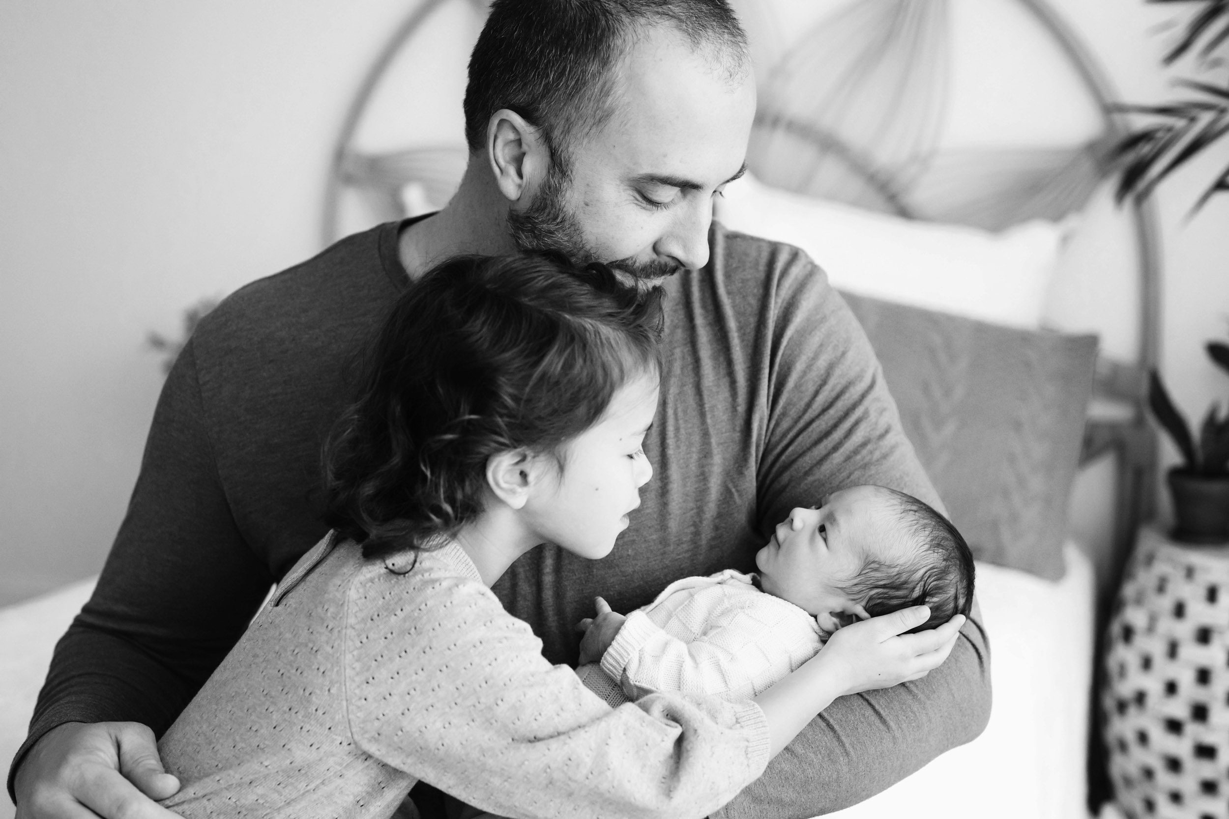 a black and white picture of a dad cradling his newborn son in his arms and hugging his older daughter as she reaches out to cradle the baby's head in her hands during a family centered newborn photo session