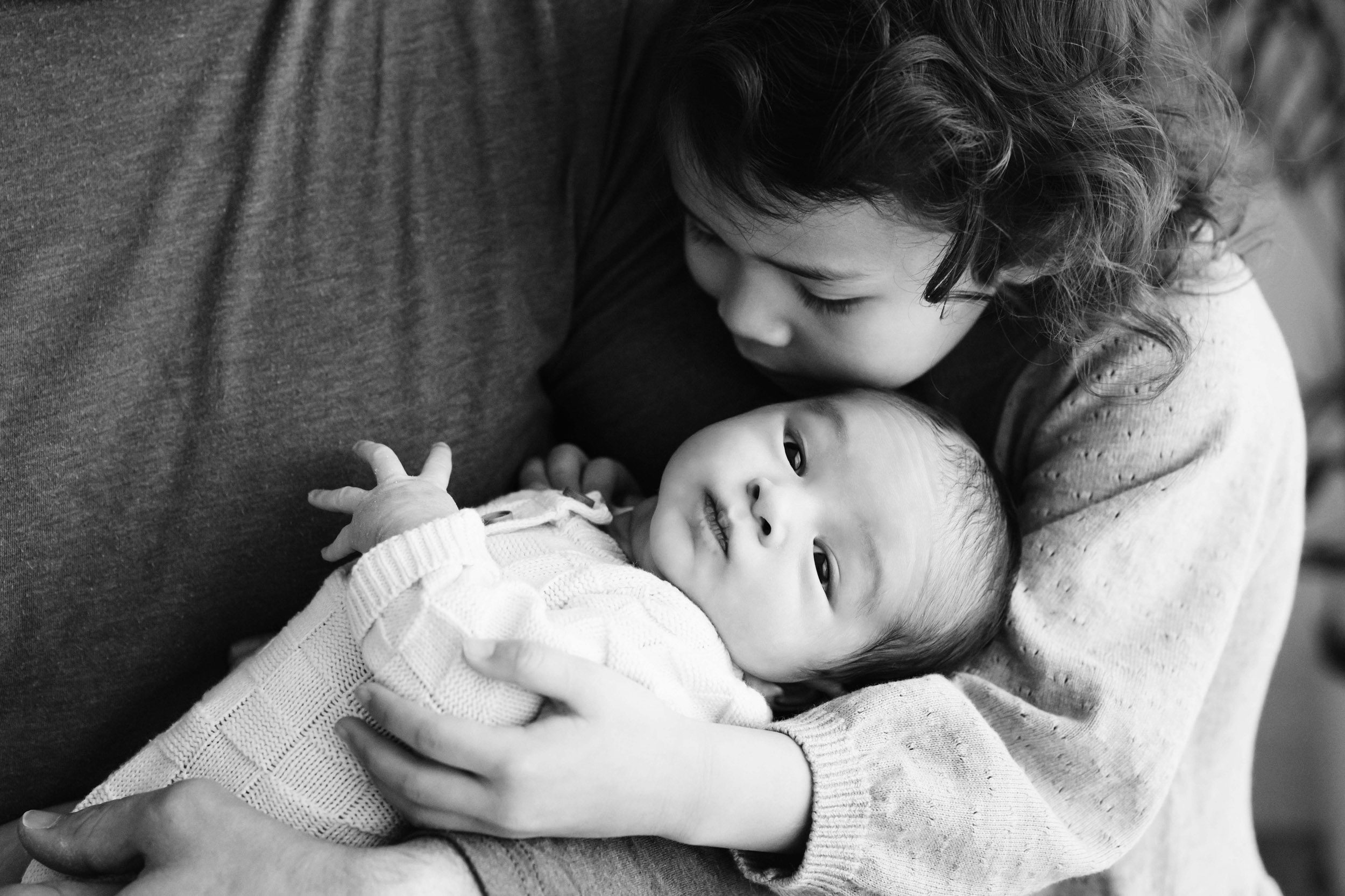 a black and white close up picture of a big sister hugging her baby brother while he's cradled in dad's arms during a family centered newborn photo session