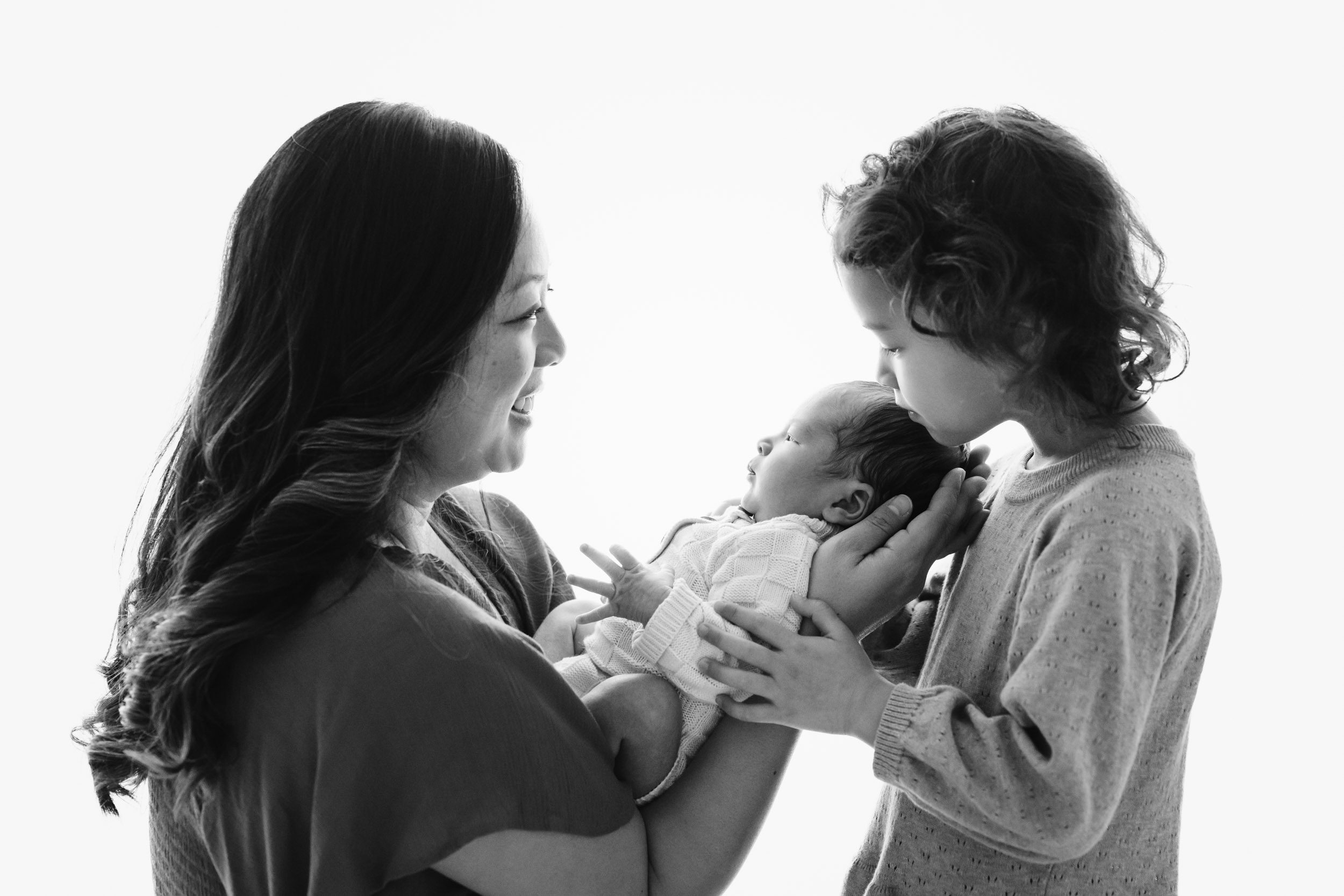 a black and white backlit photo of a mom holding her baby boy in her arms and smiling at her older daughter as she gently touches the baby during a family centered newborn photos session