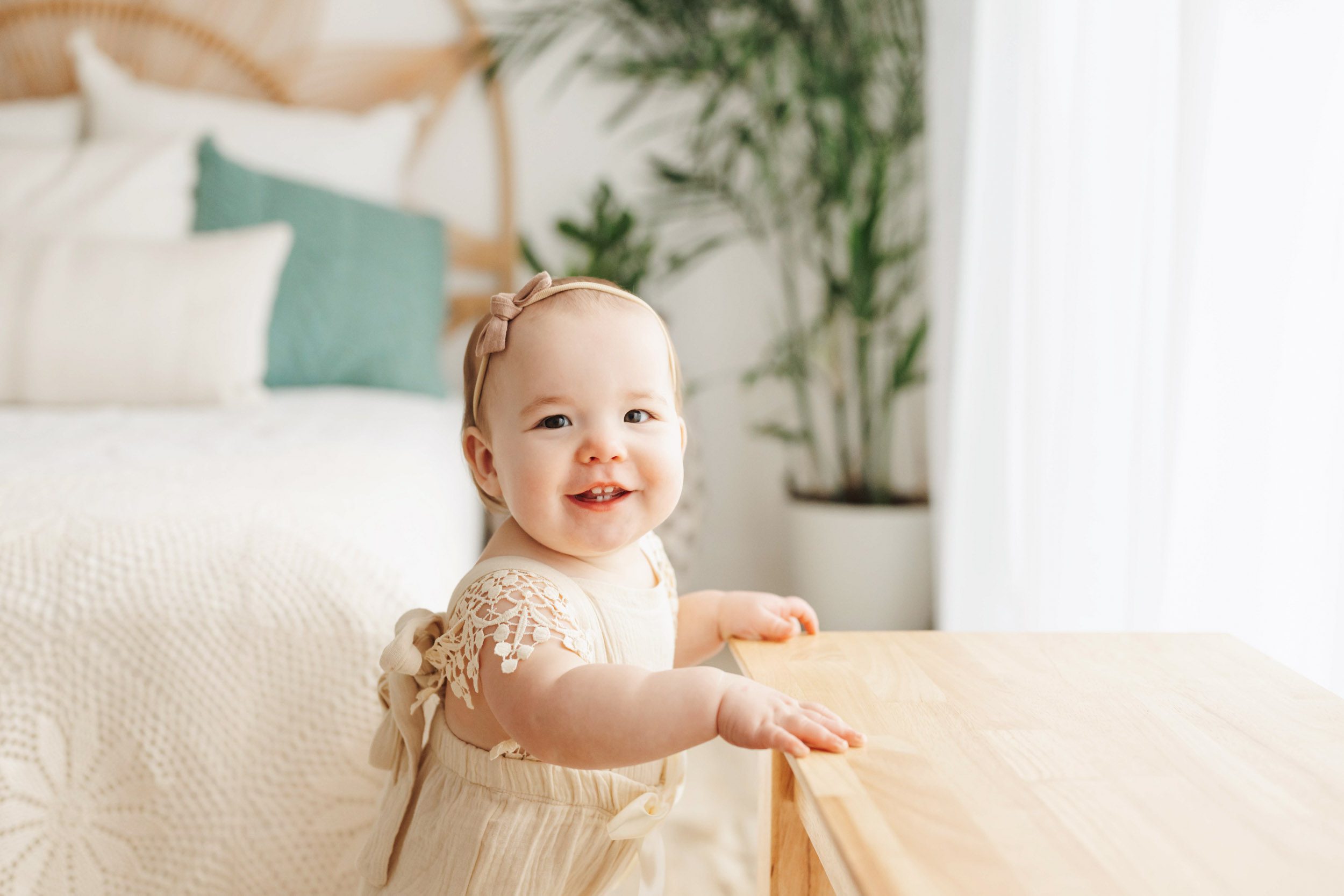a young girl standing up and holding onto a bench as she looks right at the camera with a big toothy grin on her face during a 1st birthday photos session