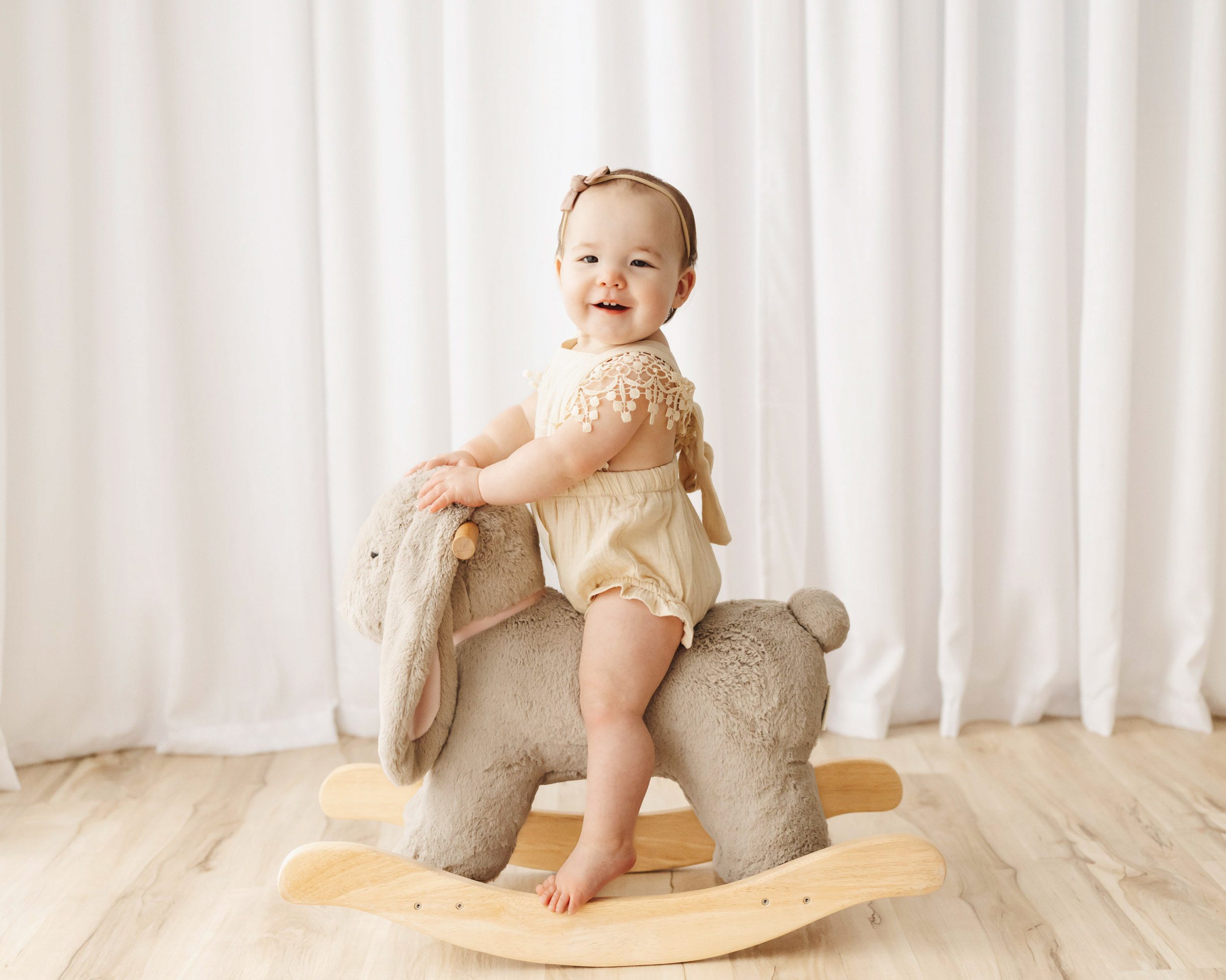 a young girl riding on a bunny rocker toy as she looks directly at the camera with a huge smile on her face during a 1st birthday photoshoot