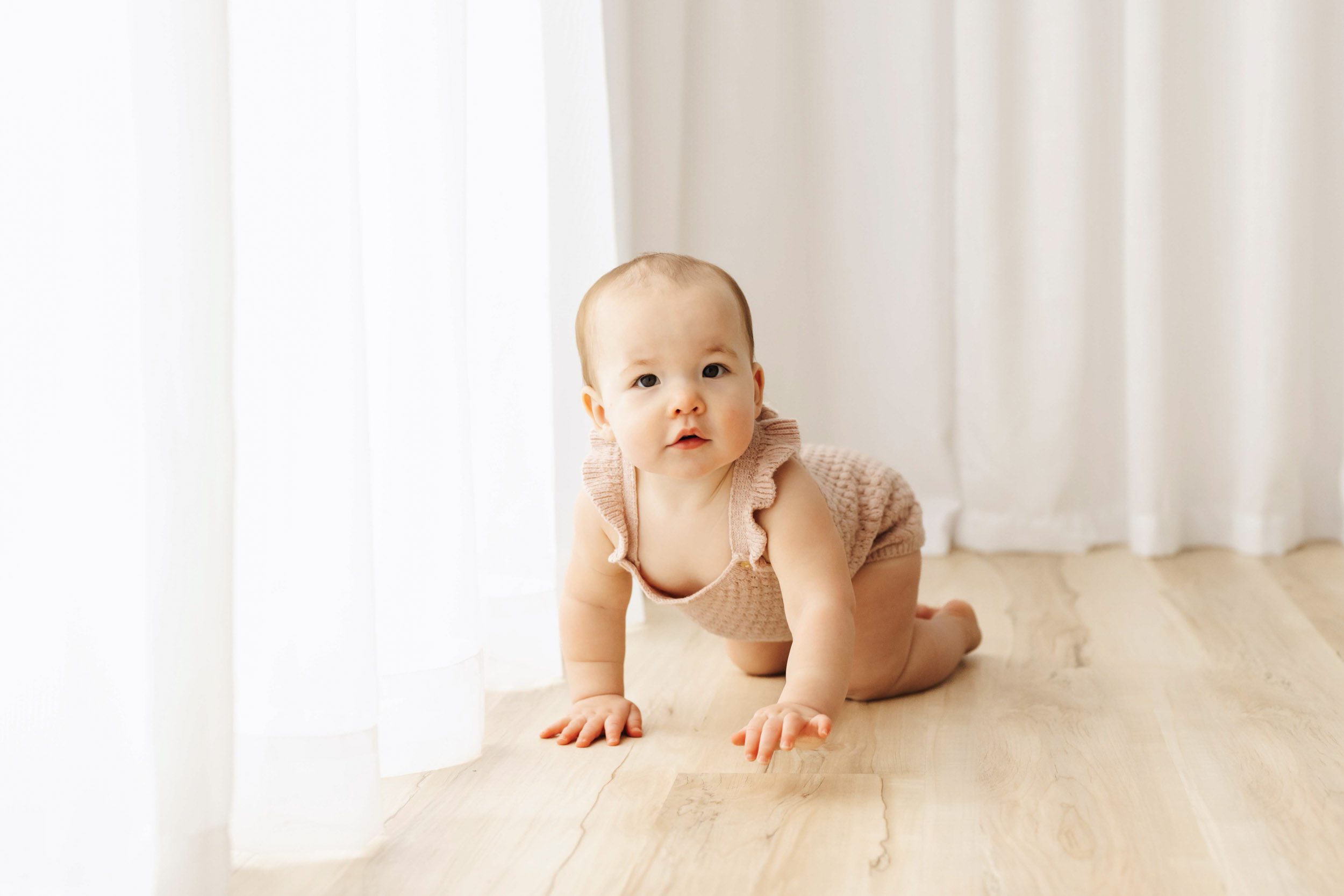 a young girl wearing a light pink knit romper crawling across the floor during a 1st birthday photoshoot