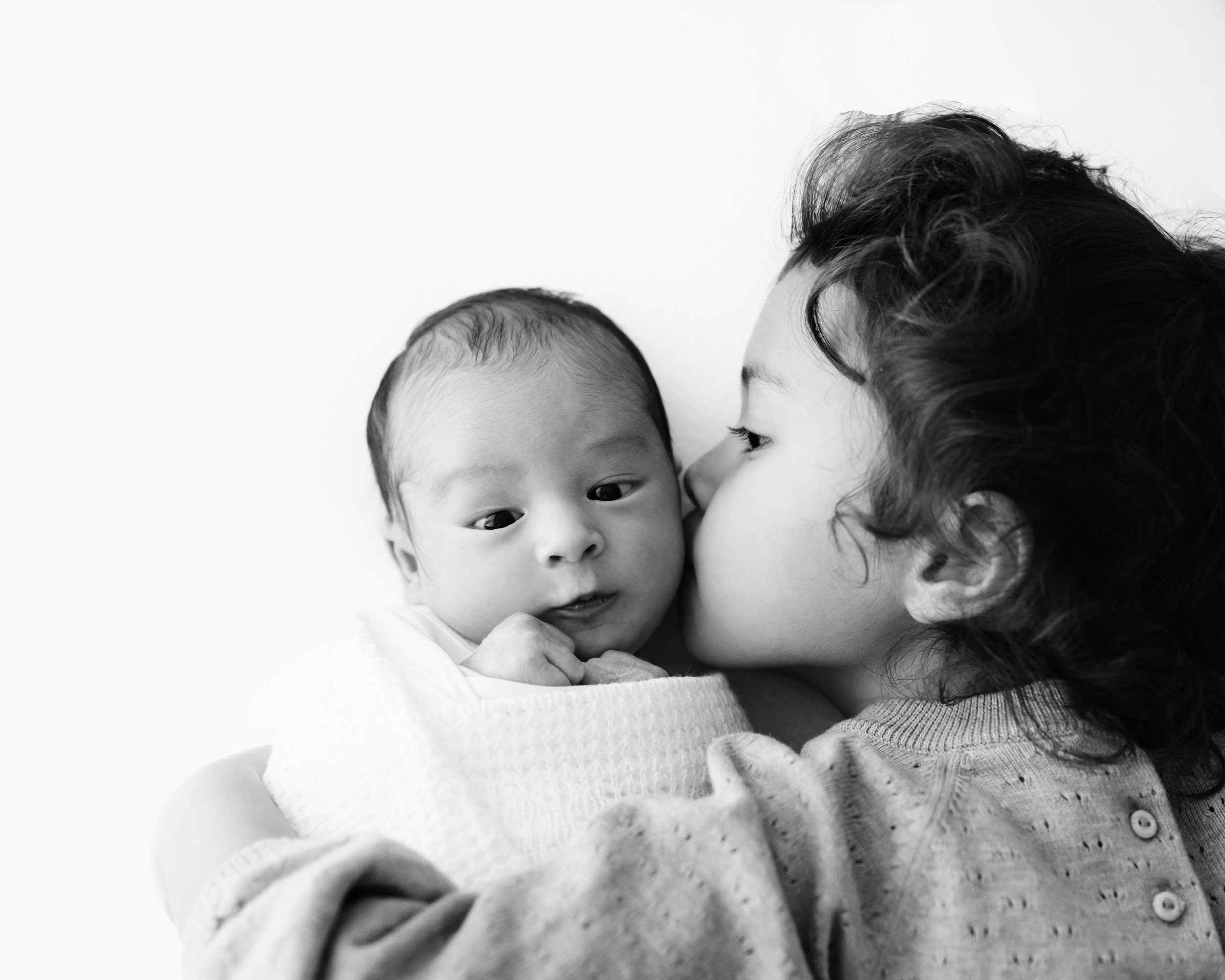 a black and white picture of a baby boy wrapped in a white swaddle blanket laying on a white backdrop as his big sister gives him a kiss on the cheek during a family centered newborn photoshoot