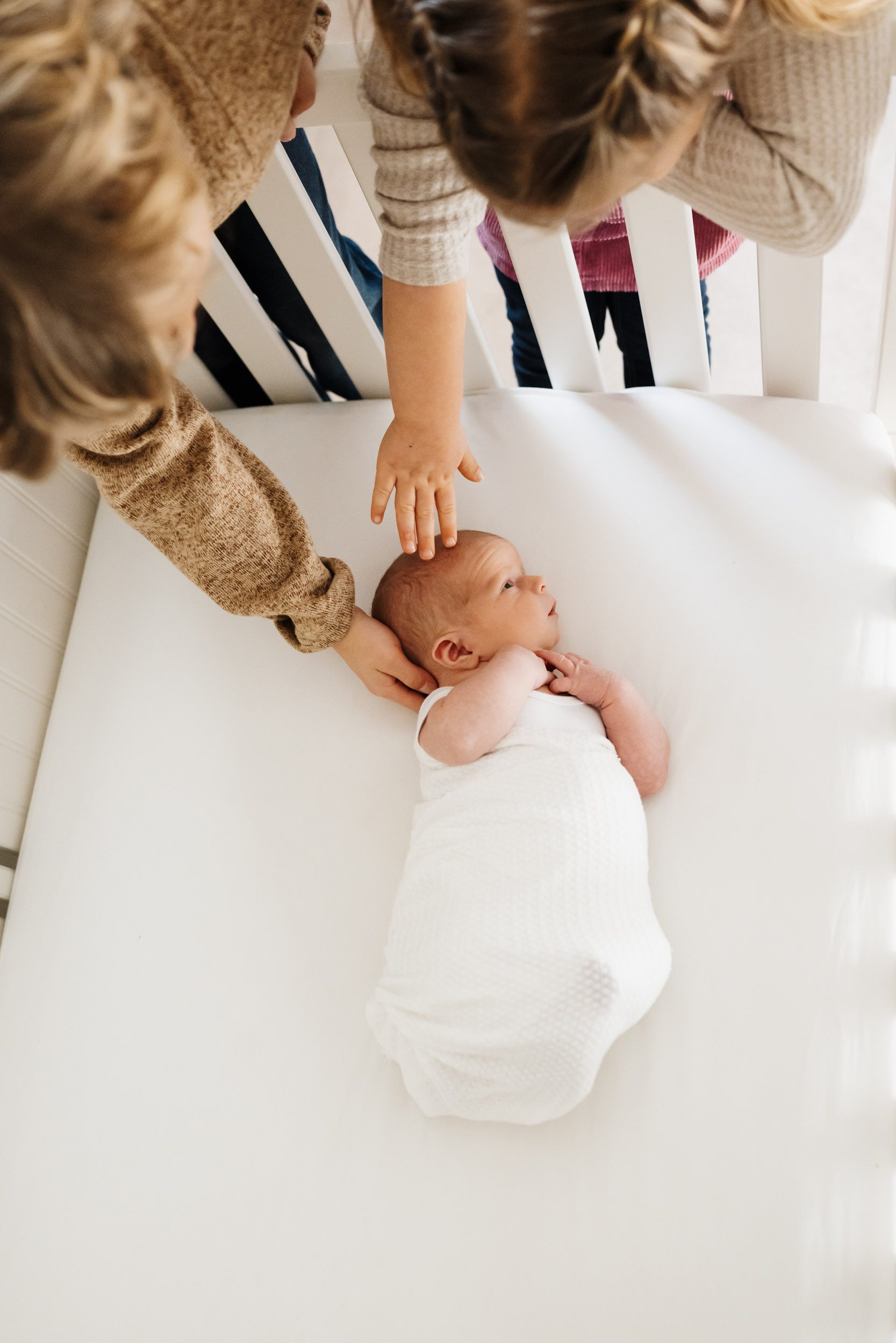 a baby boy wrapped in a white swaddle laying in his crib as his big brother and sister reach down over the edge of the crib and touch him gently on the head during an in home newborn photoshoot