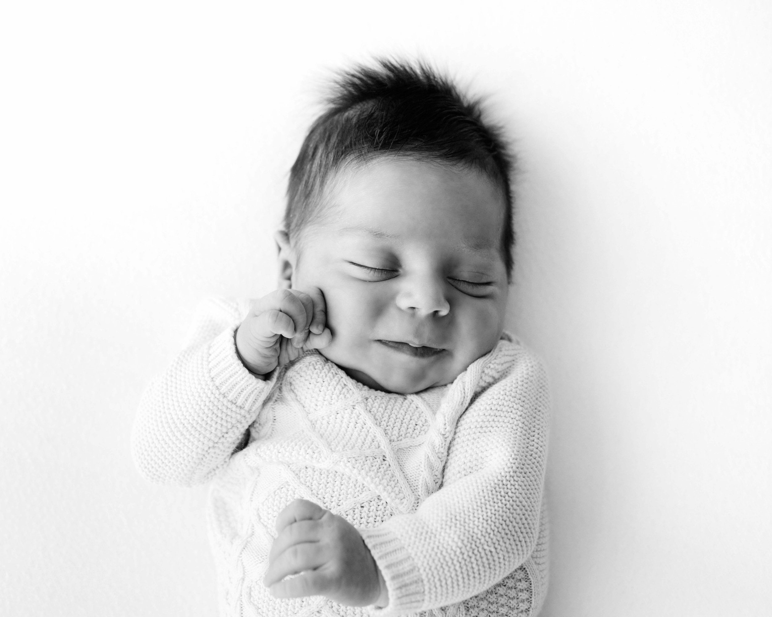 a black and white picture of a baby boy laying on a white backdrop and touching his cheek with his hand with a hint of a smile on his face during a newborn photoshoot