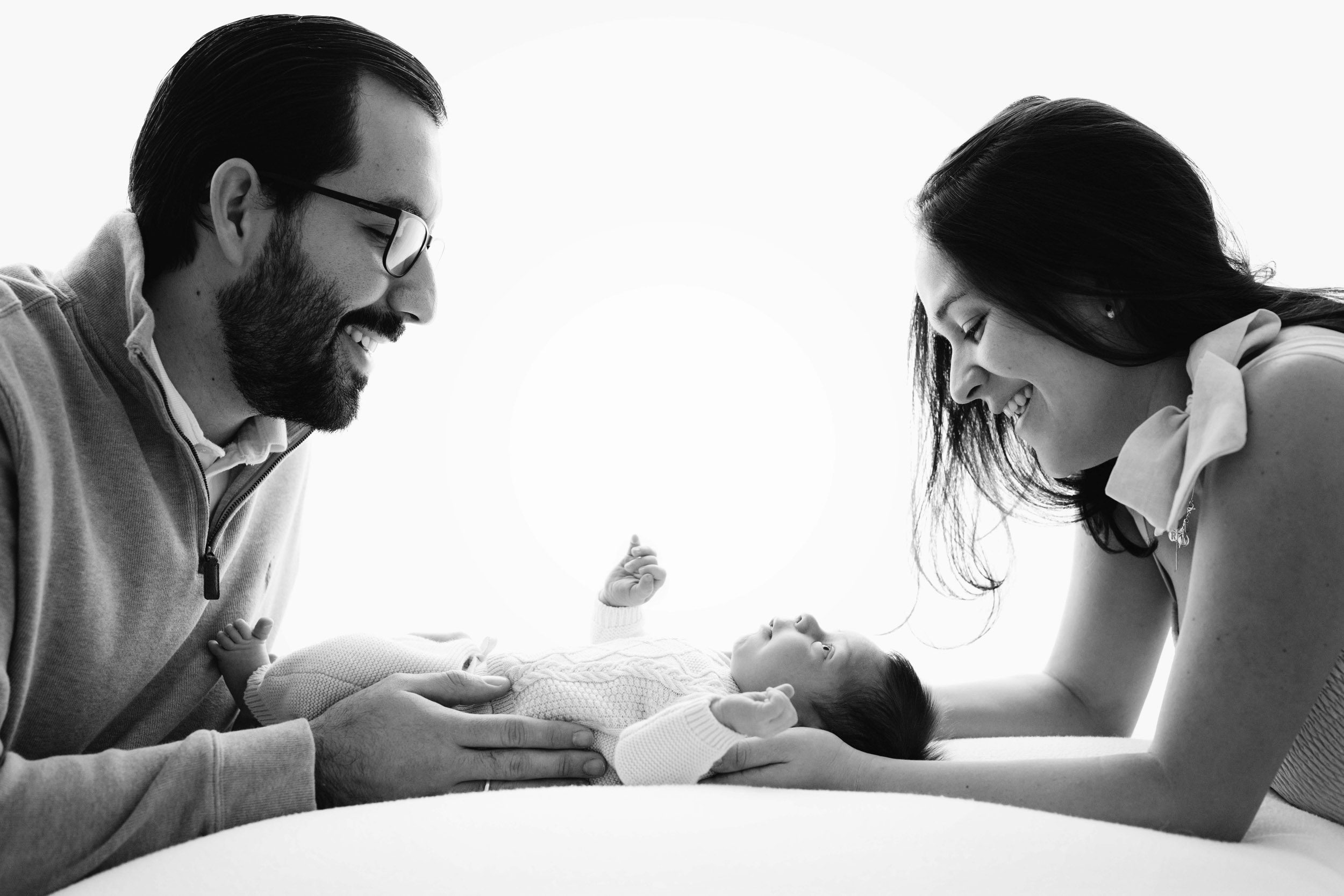 a black and white backlit picture of a baby boy laying on a white bean bag with his parents smiling down at him during a newborn photoshoot