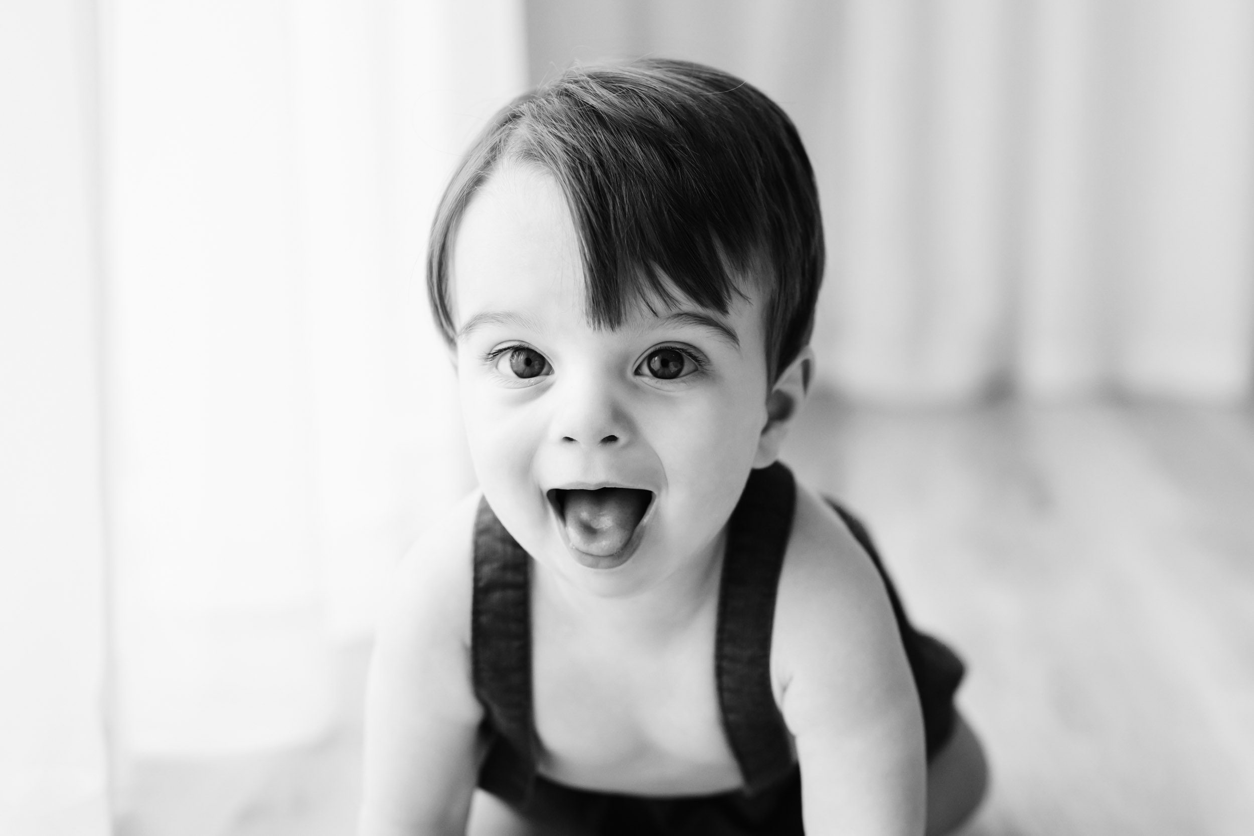 a black and white picture of a young boy crawling toward the camera and sticking out his tongue during a 1st birthday photoshoot