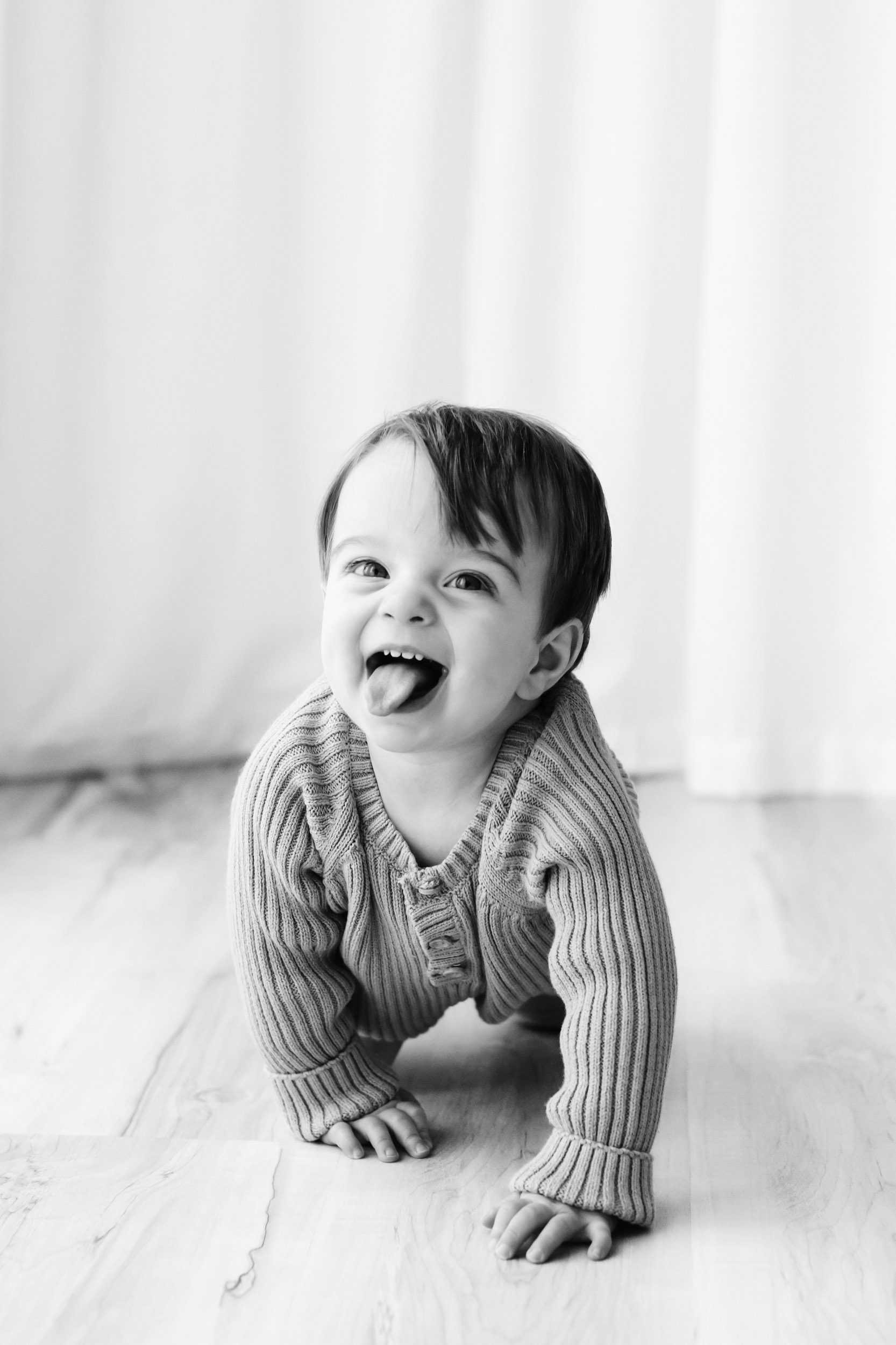 a black and white picture of a little boy crawling toward the camera with a huge smile on his face as he sticks out his tongue during a 1st birthday photoshoot