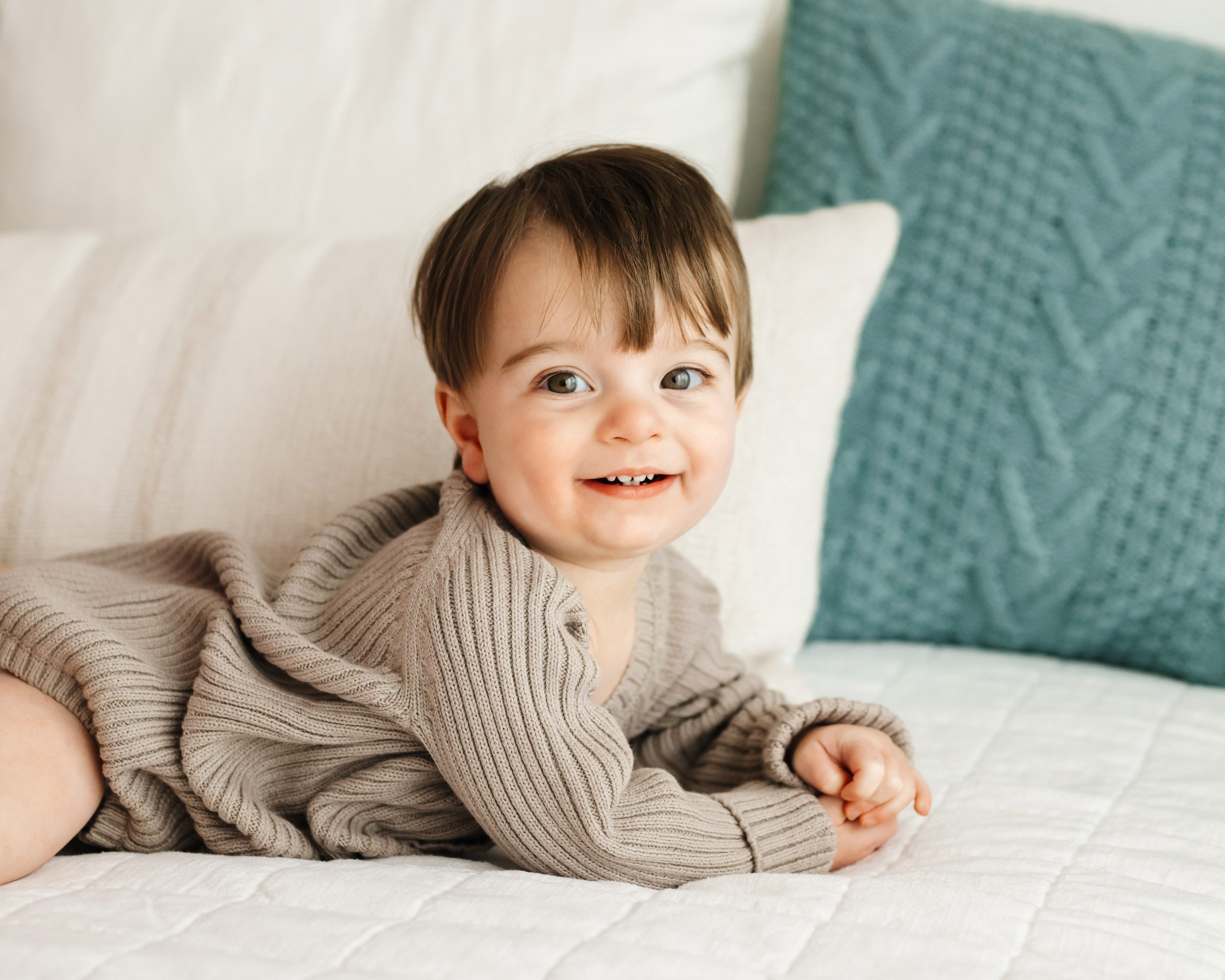 a young boy laying on his belly on a bed and smiling at the camera during a 1st birthday photoshoot