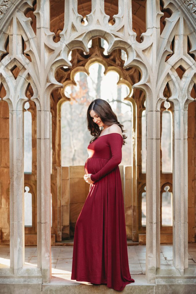 an expecting mom in a long sleeve wine colored dress standing in the middle of a stone archway and smiling down at her belly during a winter maternity photoshoot