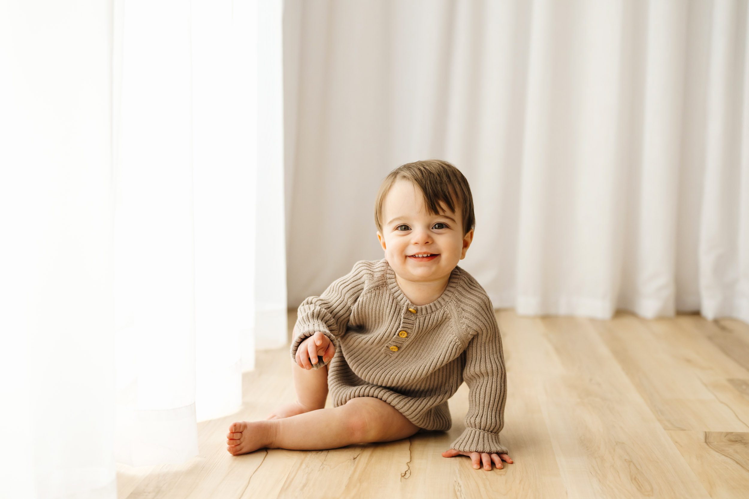 a young boy sitting on the floor next to a wall of white curtains and smiling at the camera during a 1st birthday photoshoot