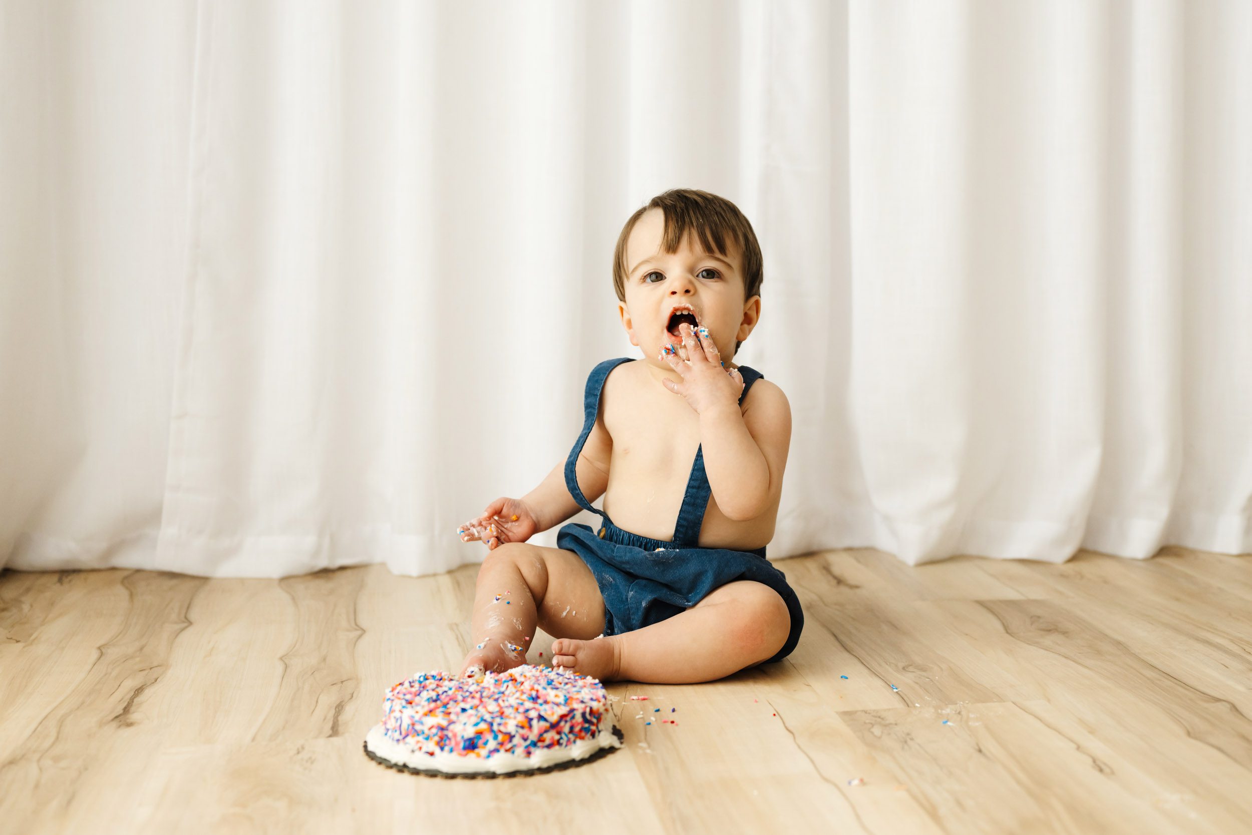 a young boy sitting the floor in front of a wall of white curtains and eating a cake covered with colorful sprinkles during a 1st birthday cake smash photoshoot