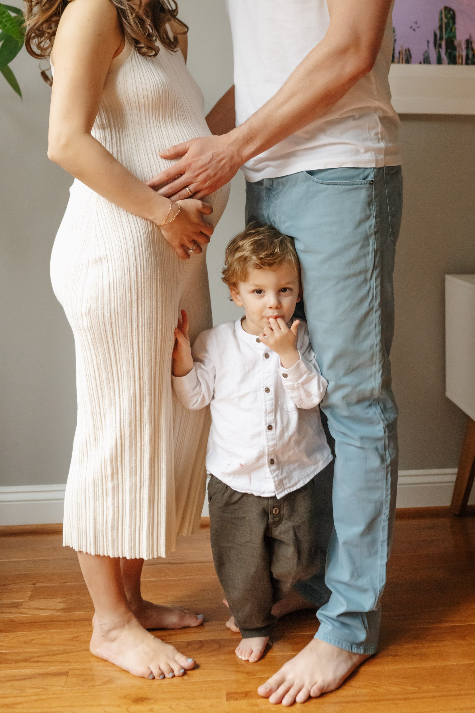 a close up picture of a little boy looking right at the camera and sucking on his fingers while his mom and dad stand next to him and both touch mom's belly during an in home maternity photoshoot