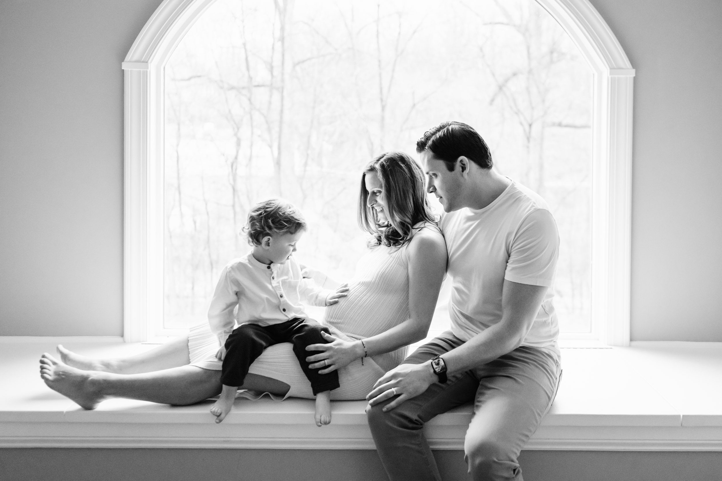 a black and white backlit image of a family sitting in front of a bay window with their son gently touching his mom's belly during an in home maternity photoshoot