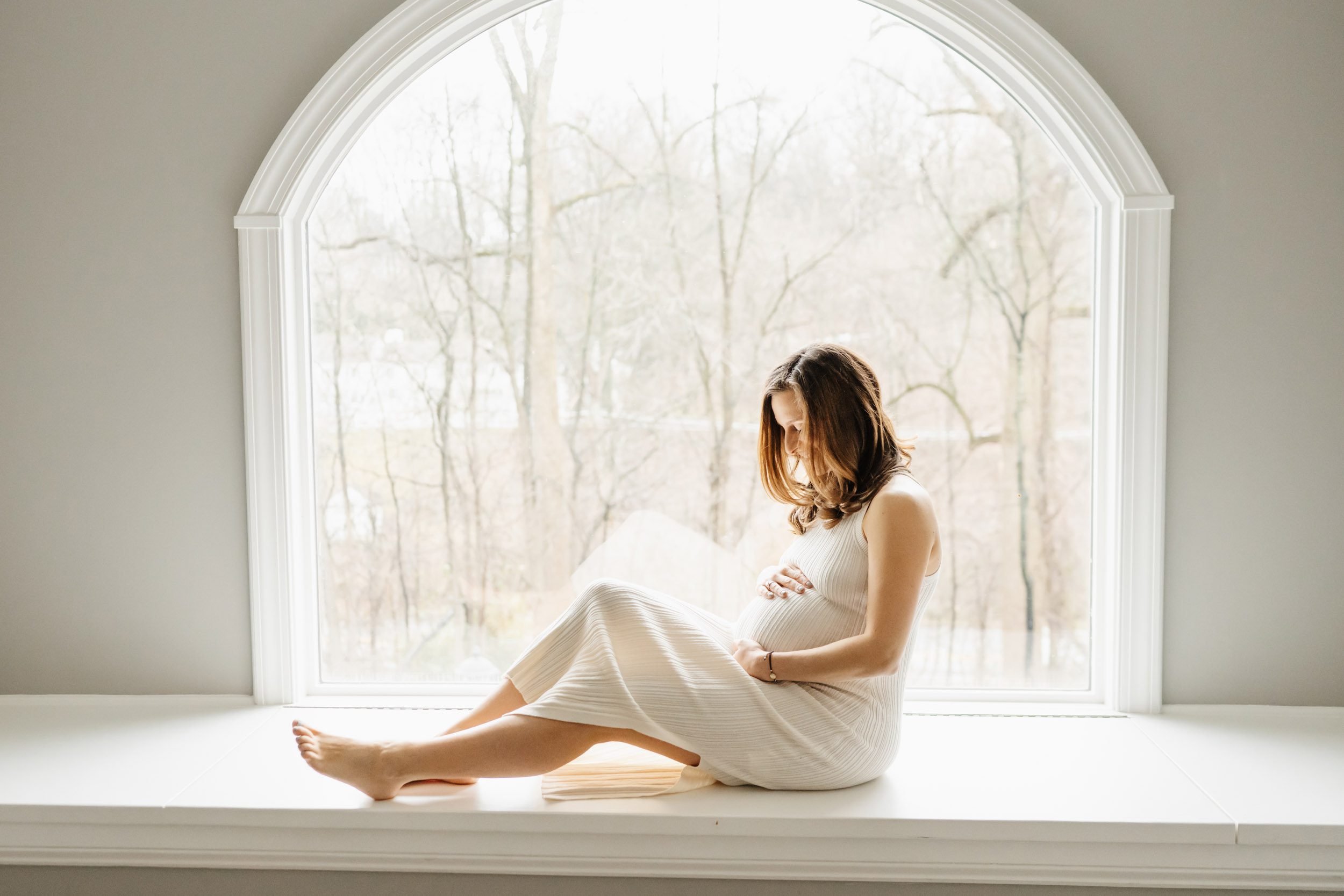 a backlit picture of an expecting mom sitting in front of a bay window and cradling her belly in her hands during an in home maternity photoshoot