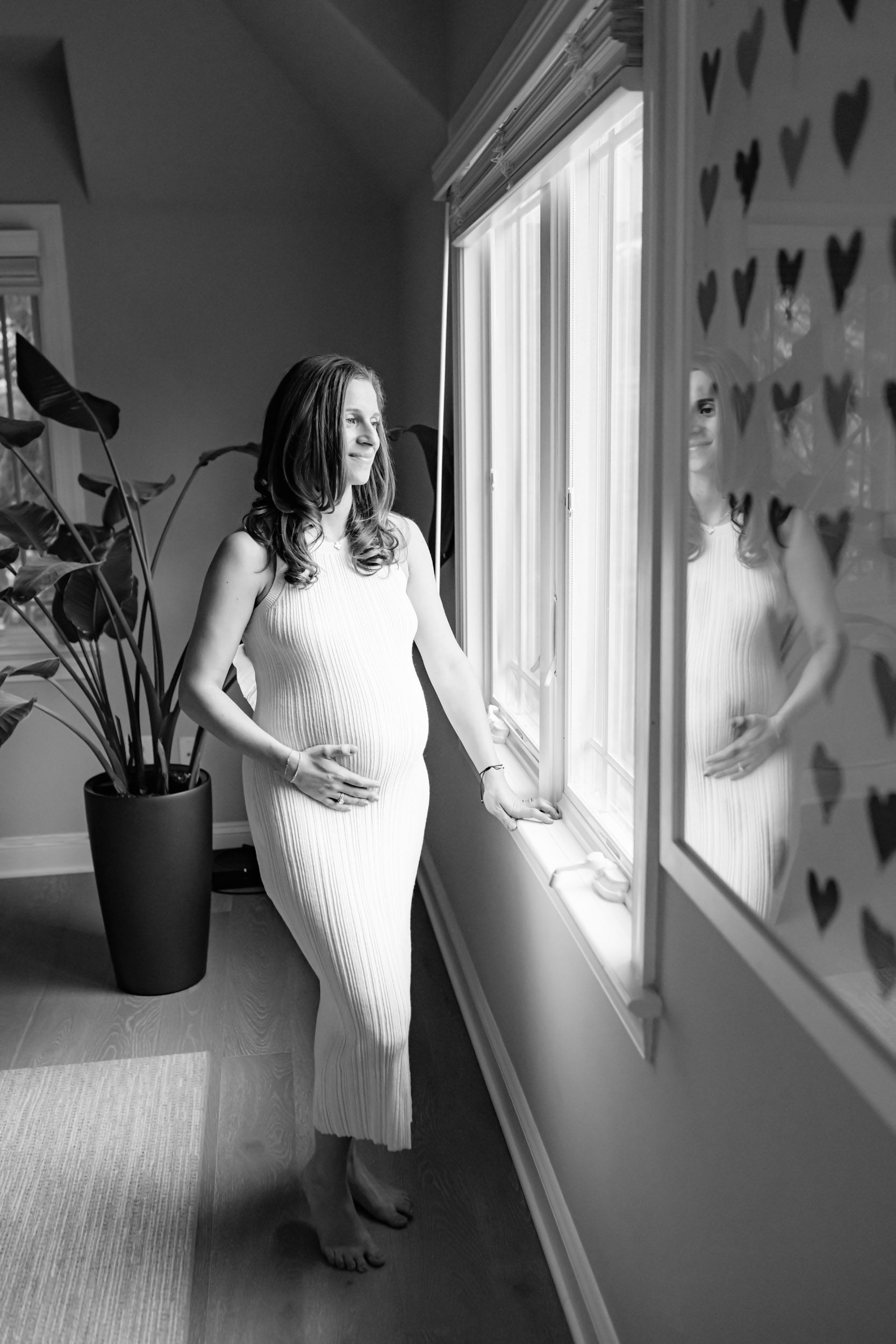 a black and white picture of an expecting mom standing next to a window and touching her belly as she gazes out the window during an in home maternity photoshoot