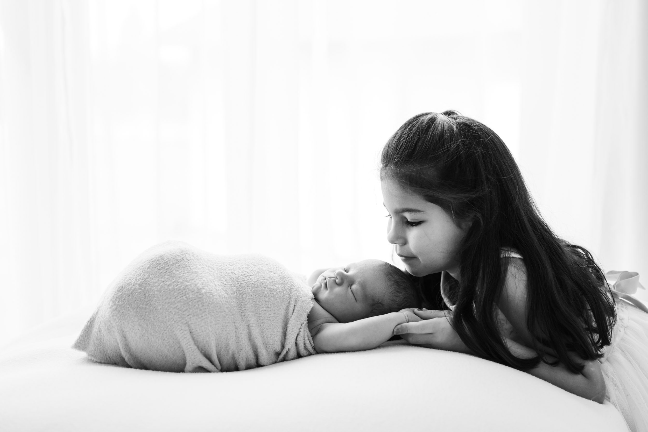 a black and white backlit photo of a baby boy laying on a bean bag in front of a wall of windows while his young sibling holds his hands and smiles down at him during a newborn photos session