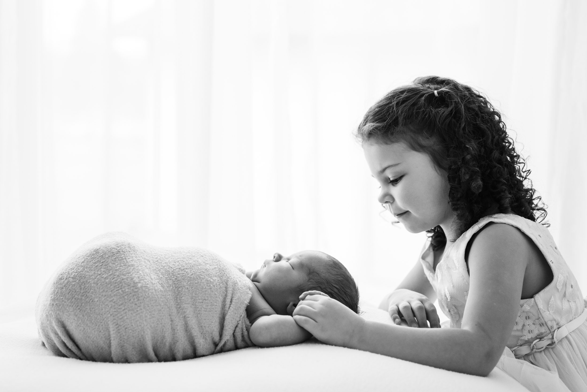 a black and white backlit photo of a baby boy laying on a bean bag in front of a wall of windows while his young sibling holds his hand and smiles down at him during a newborn photos session