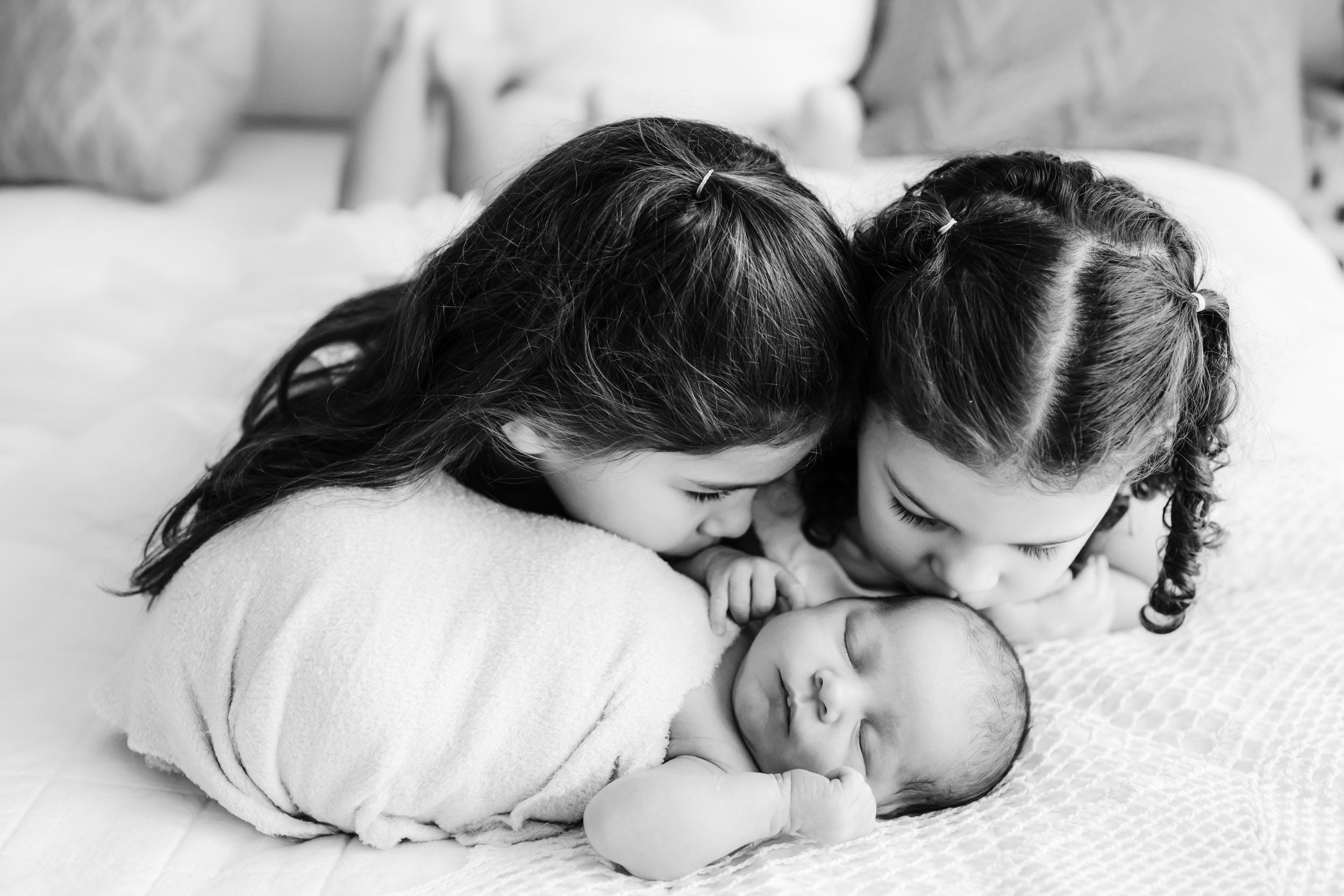 a black and white picture of young siblings laying with their baby brother on a bed as the both kiss him during a newborn photos session