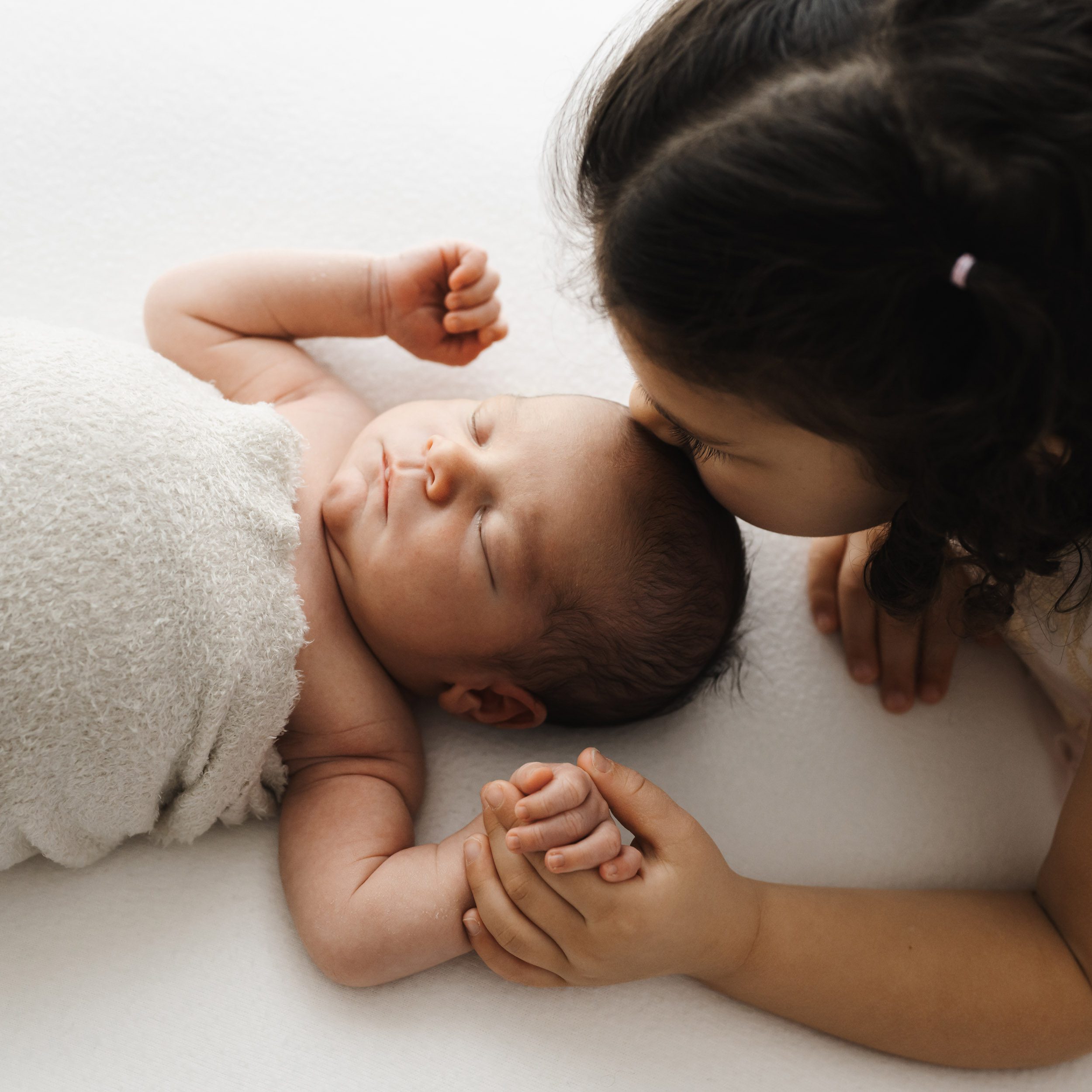a picture taken from above of a baby boy laying on a beanbag as his young sibling holds his hand and kisses him on the head during a newborn photos session