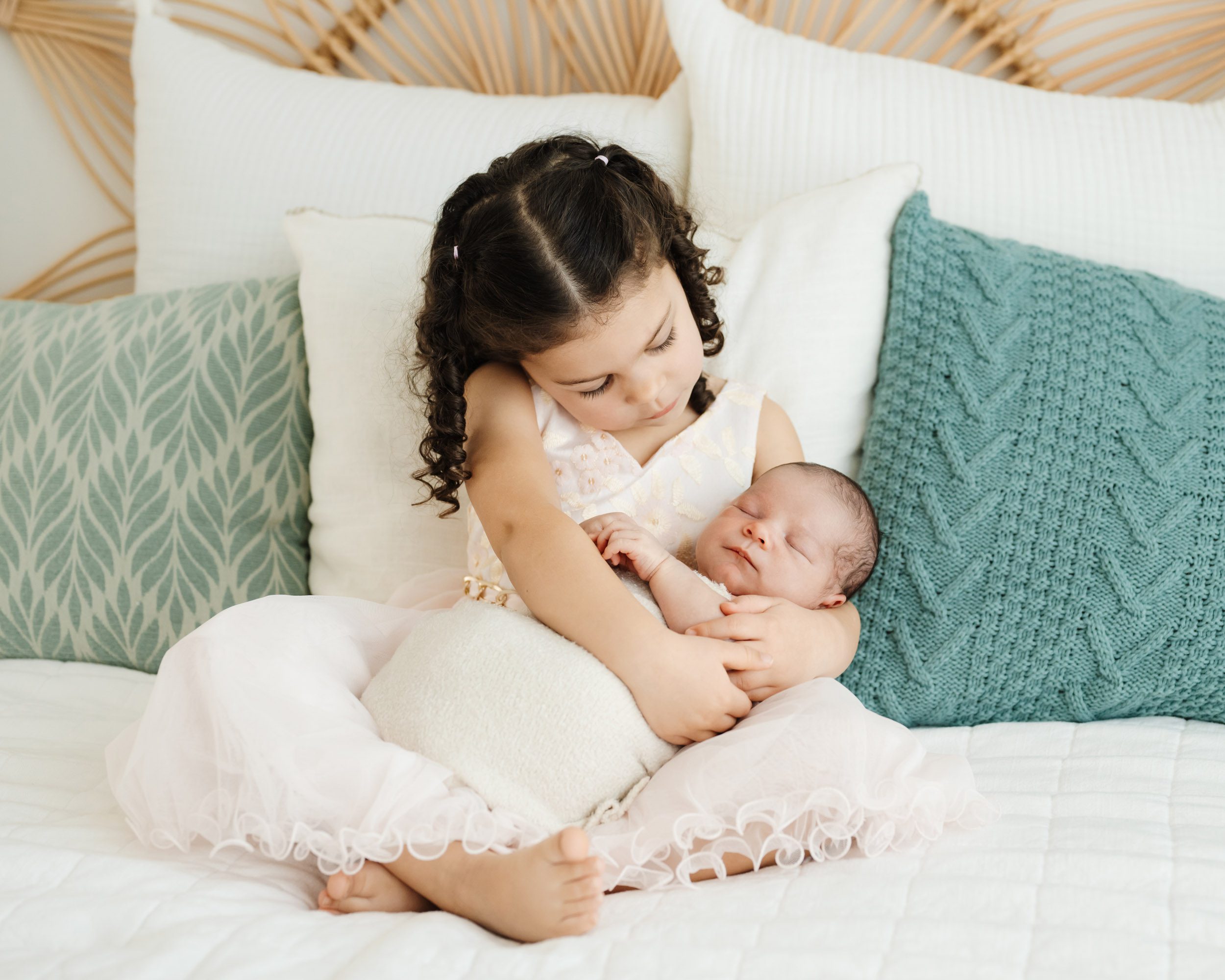 a young sibling sitting on a bed and cradling her baby brother in her arms as she smiles down at him during a newborn photos session