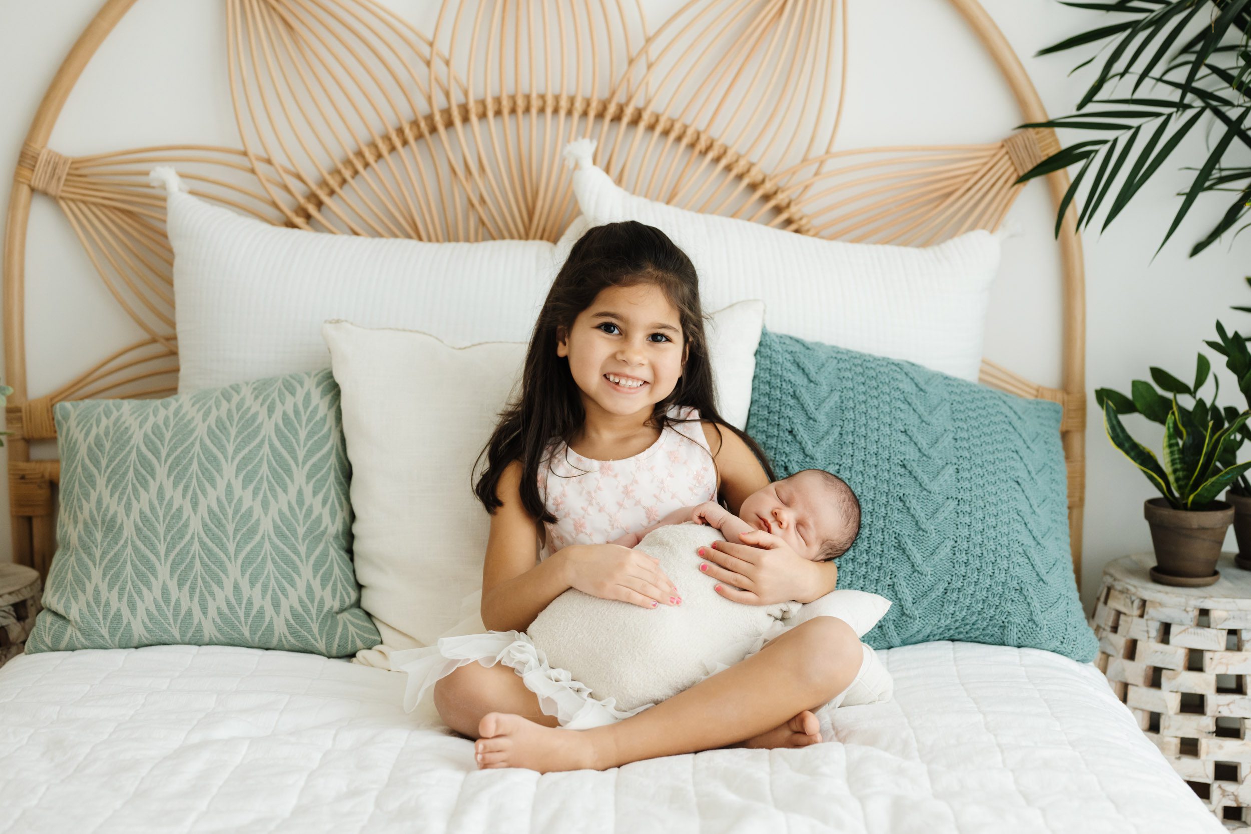 a young sibling sitting on a bed and cradling her baby brother in her arms as she smiles at the camera during a newborn photo session