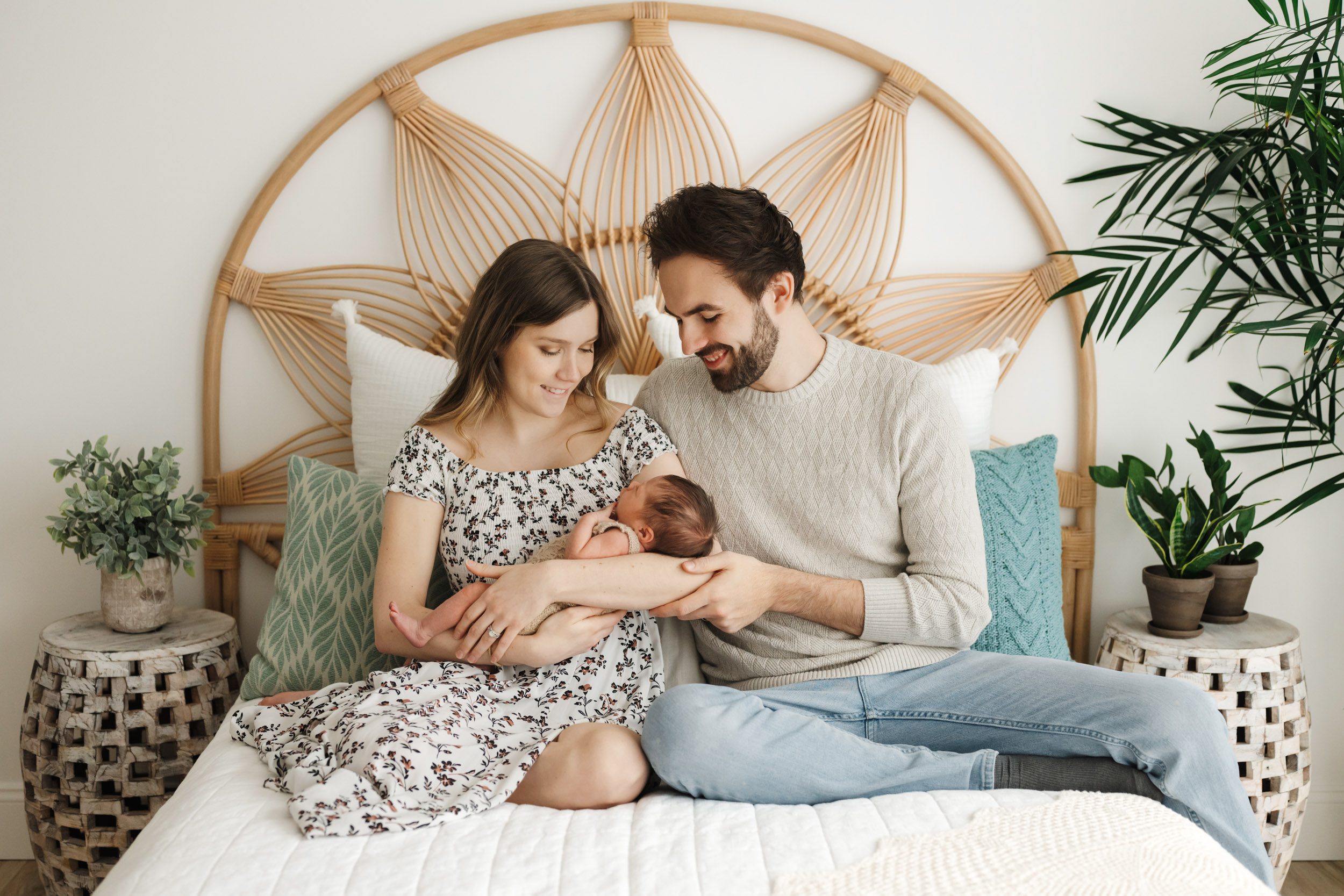 new parents sitting on a bed holding their baby girl in their arms and smiling down at her during a newborn photos session