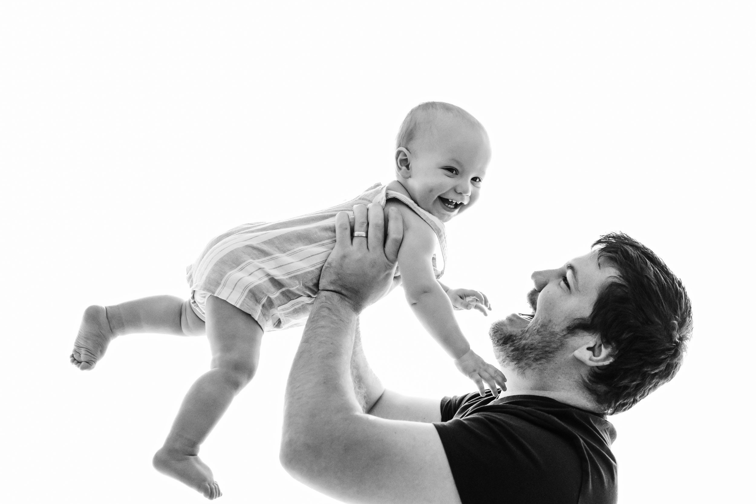 a black and white backlit picture of a little boy smiling as his dad flies him up in the air during a baby's 1st year photoshoot