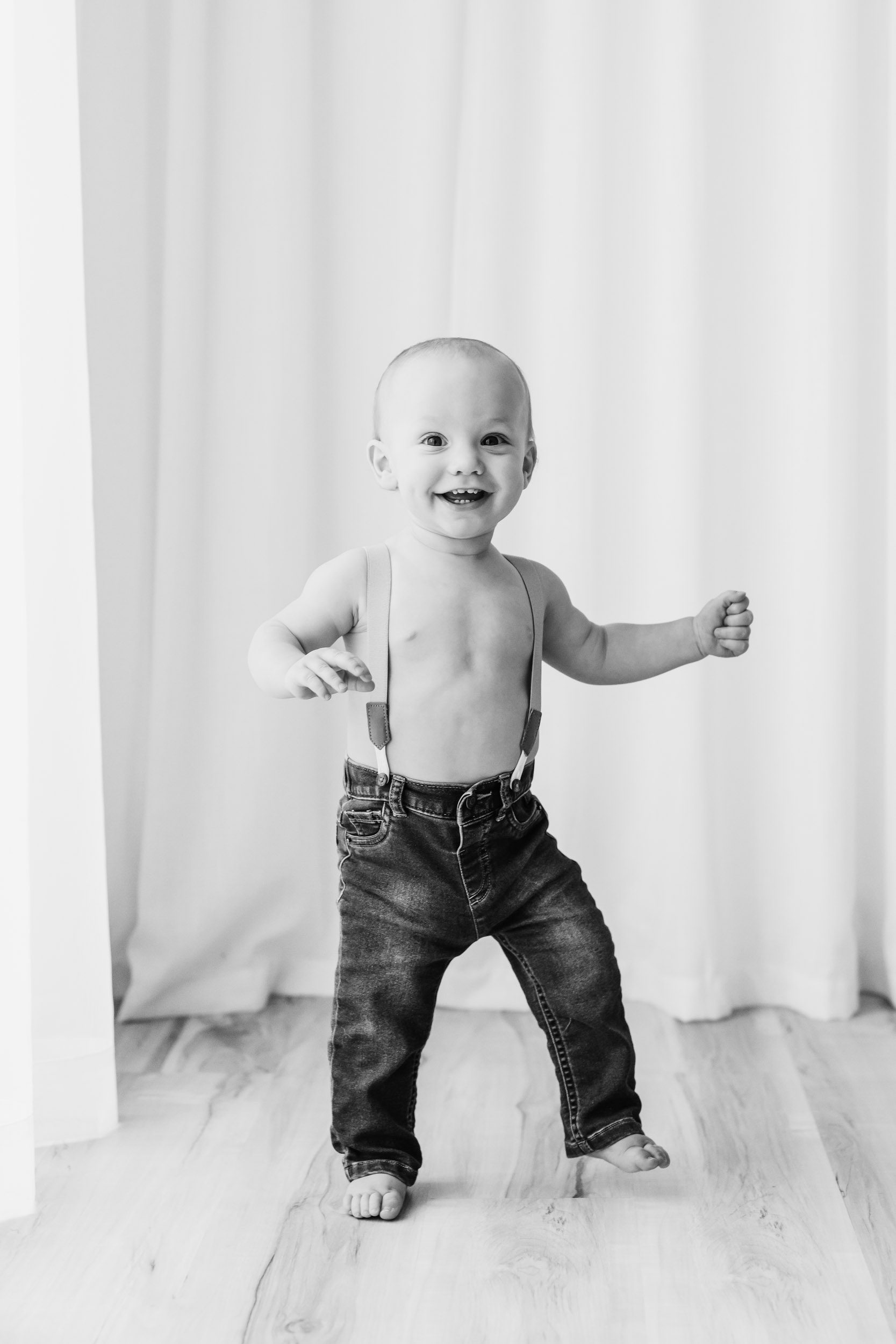a black and white picture of a young boy wearing jeans and suspenders walking toward the camera with a smile on his face during a baby's 1st year photoshoot