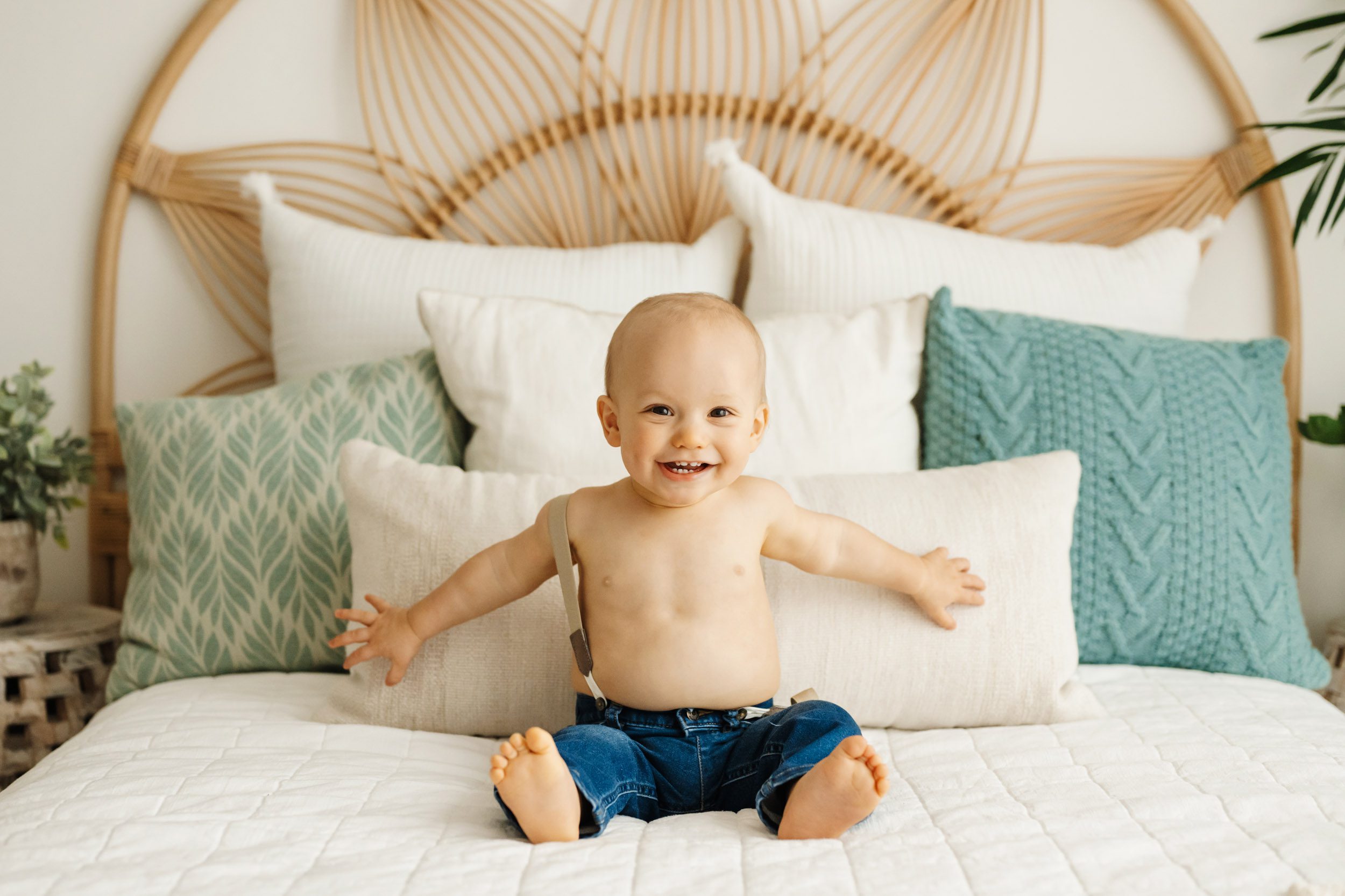 a young boy wearing blue jeans and suspenders sitting on a bed and holding his arms out wide as he smiles directly at the camera during a baby's 1st year photoshoot