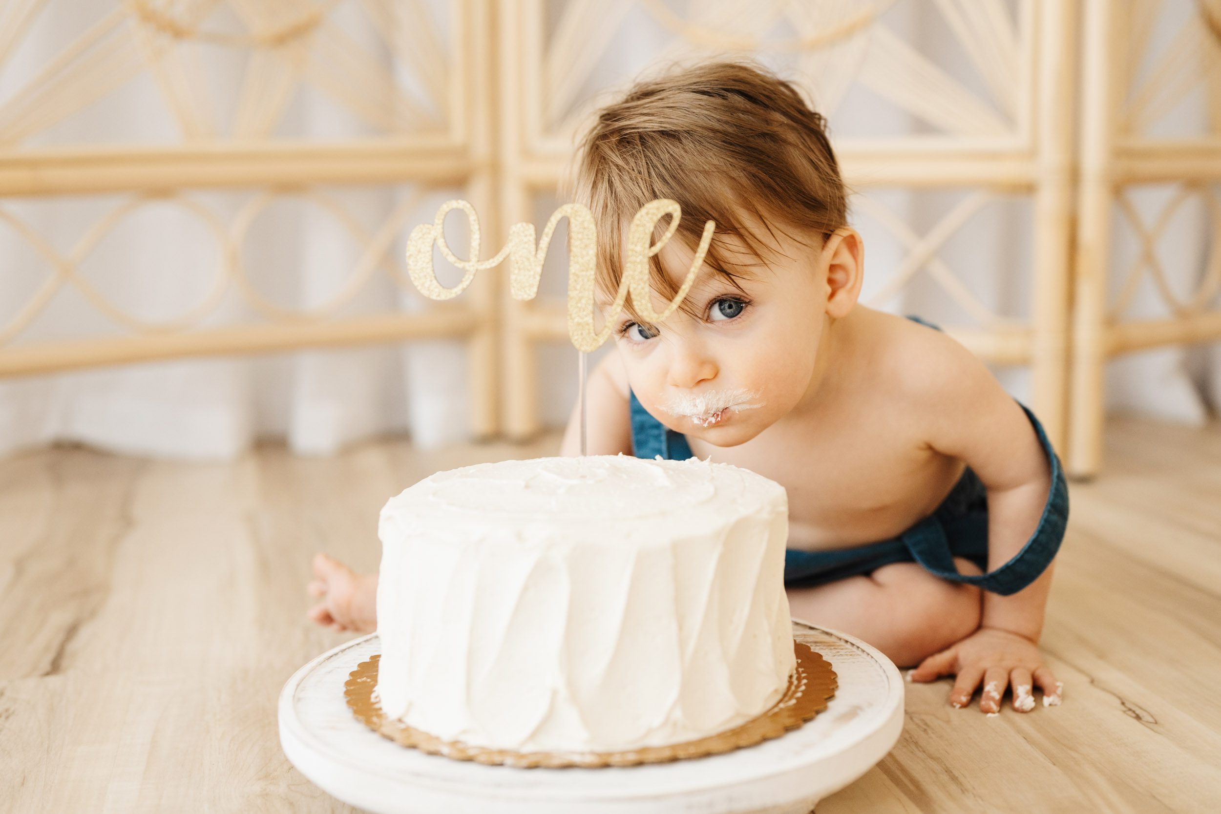 a young boy sitting on the floor and leaning forward to take a bite out of a white cake with white icing all around his mouth during a 1st birthday cake smash & splash photoshoot