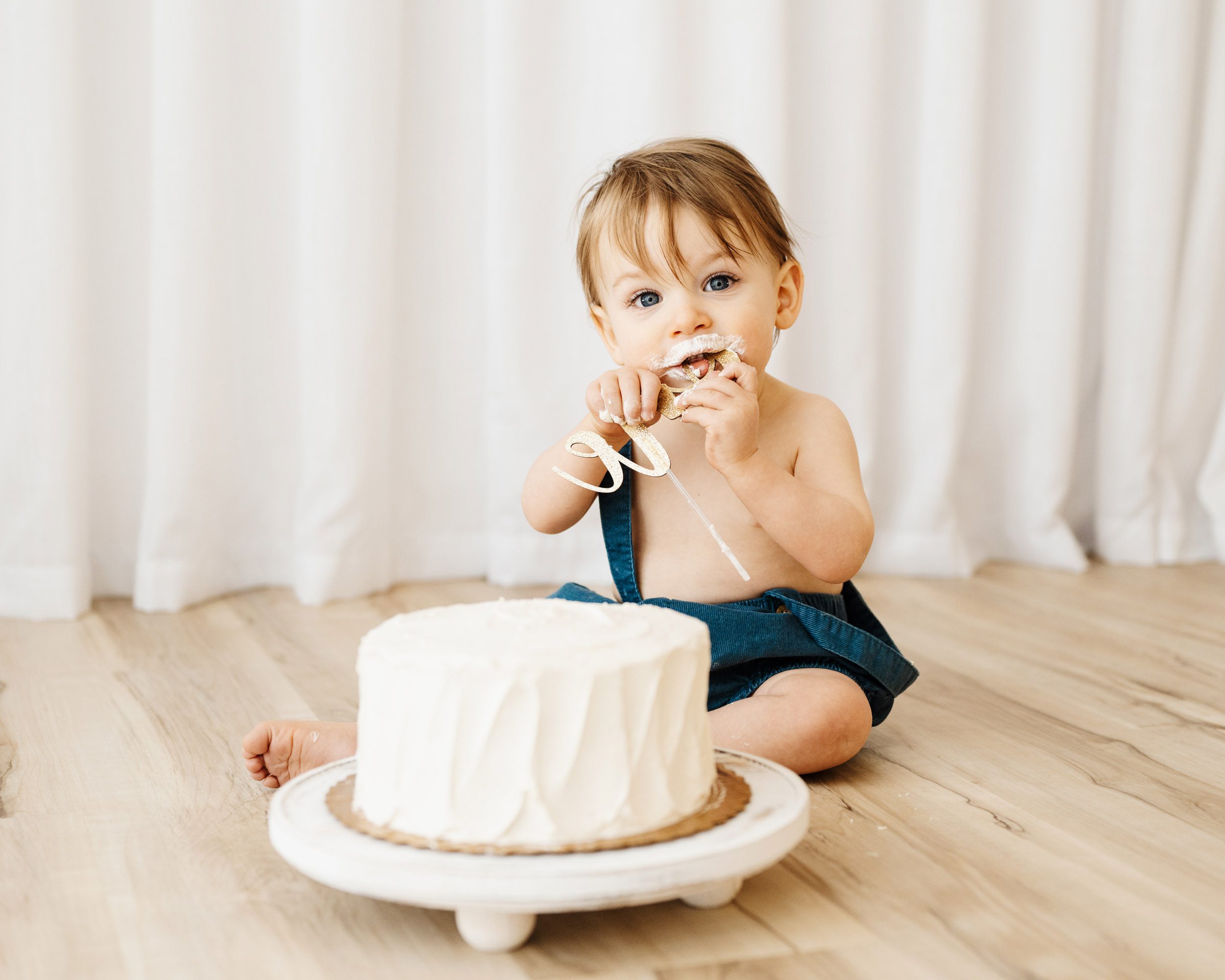 a young boy sitting on the floor next to a white cake as he chews on the cake topper decoration and looks directly at the camera during a 1st birthday cake smash & splash photoshoot