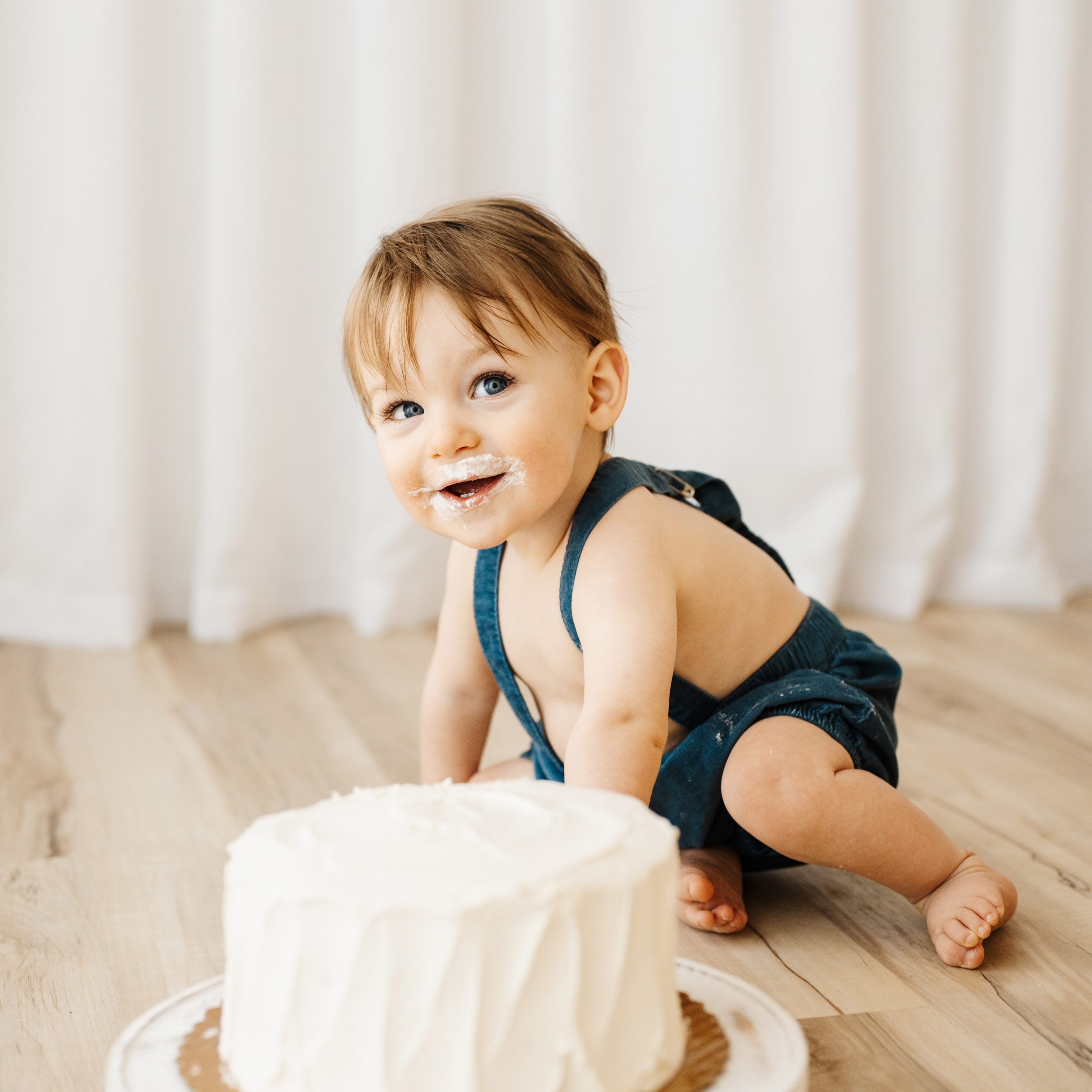 a young boy sitting on the floor behind a white cake and looking up at his mom behind the camera with a huge smile on his face during a 1st birthday cake smash & splash photoshoot