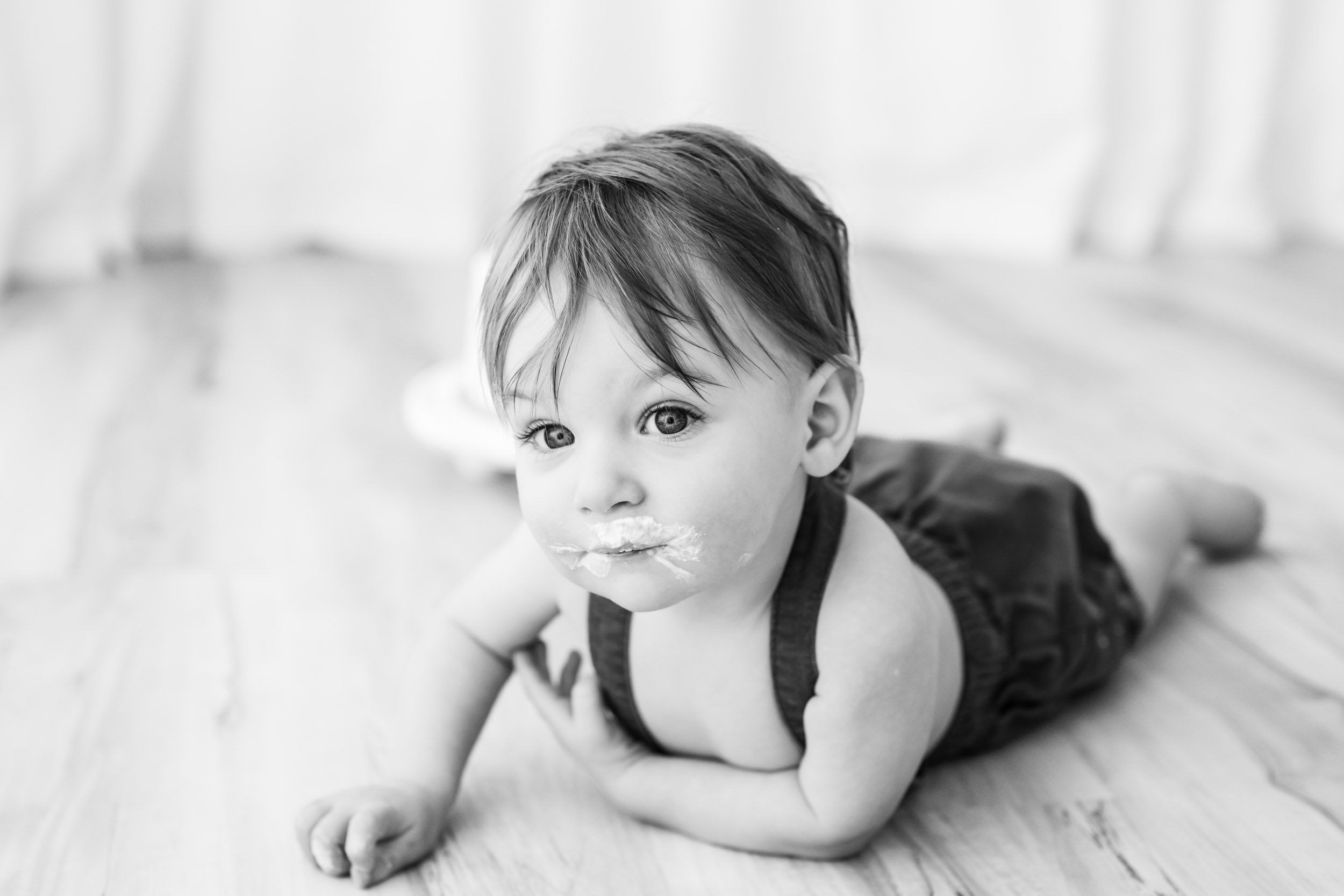 a black and white picture of a young boy crawling across the floor toward the camera and looking directly at the camera during a 1st birthday cake smash & splash photoshoot