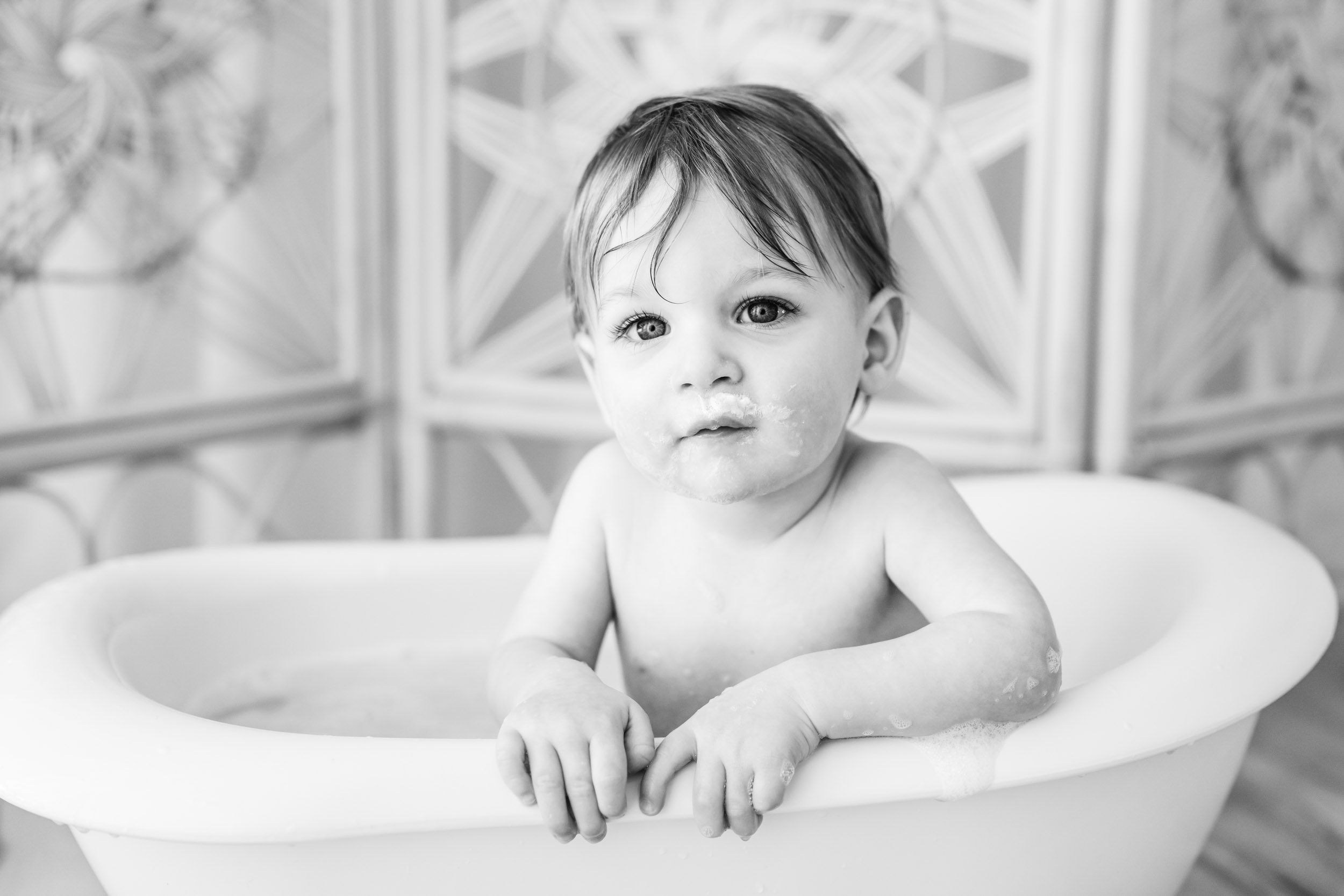 a black and white picture of a young boy sitting in a little white bath tub as he leans on the side of the tub and gazes directly into the camera a young boy splashing in a little bathtub and looking right at the camera with a huge smile on his face during a 1st birthday cake smash & splash photoshoot