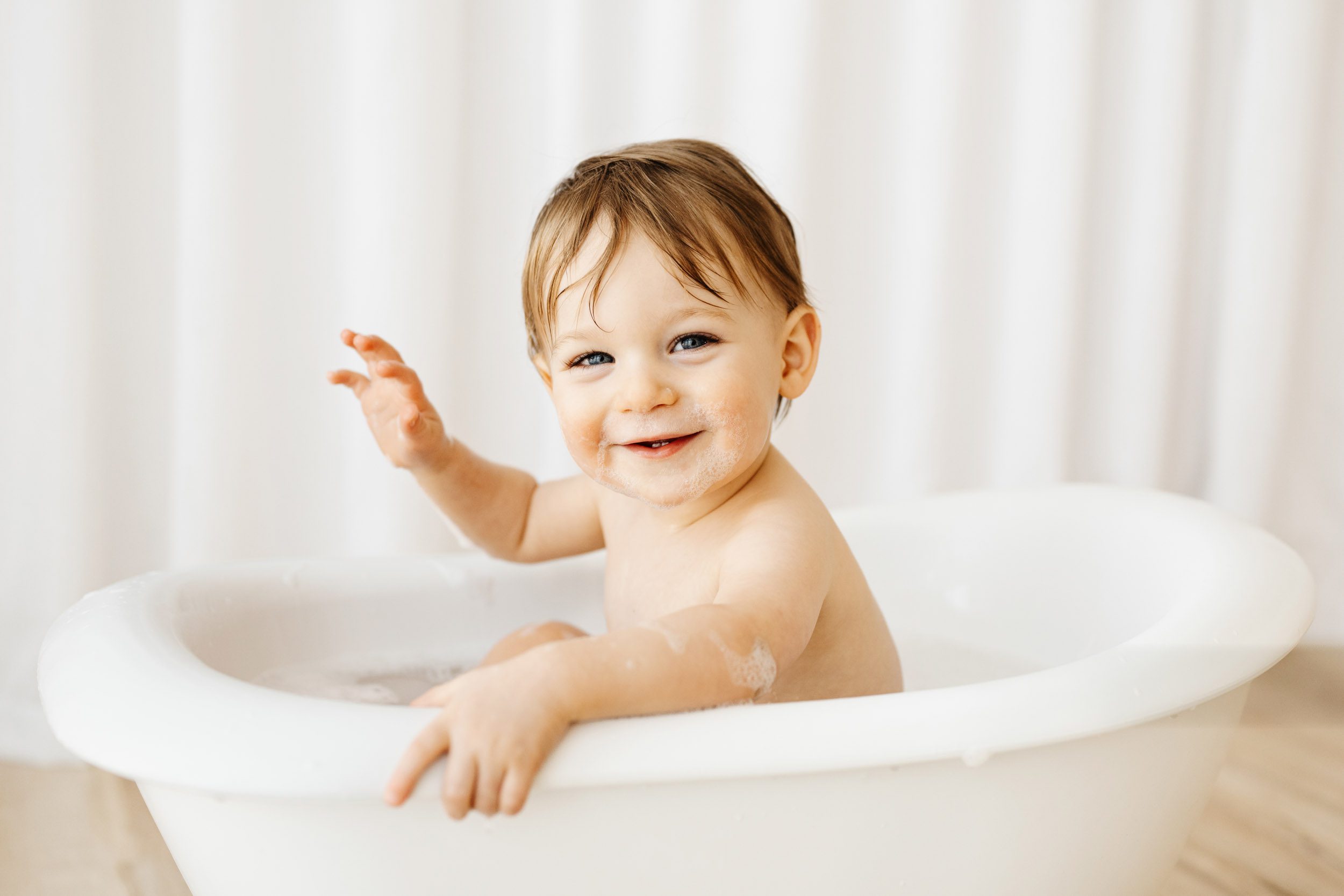 a young boy splashing in a little bathtub and looking right at the camera with a huge smile on his face during a 1st birthday cake smash & splash photoshoot