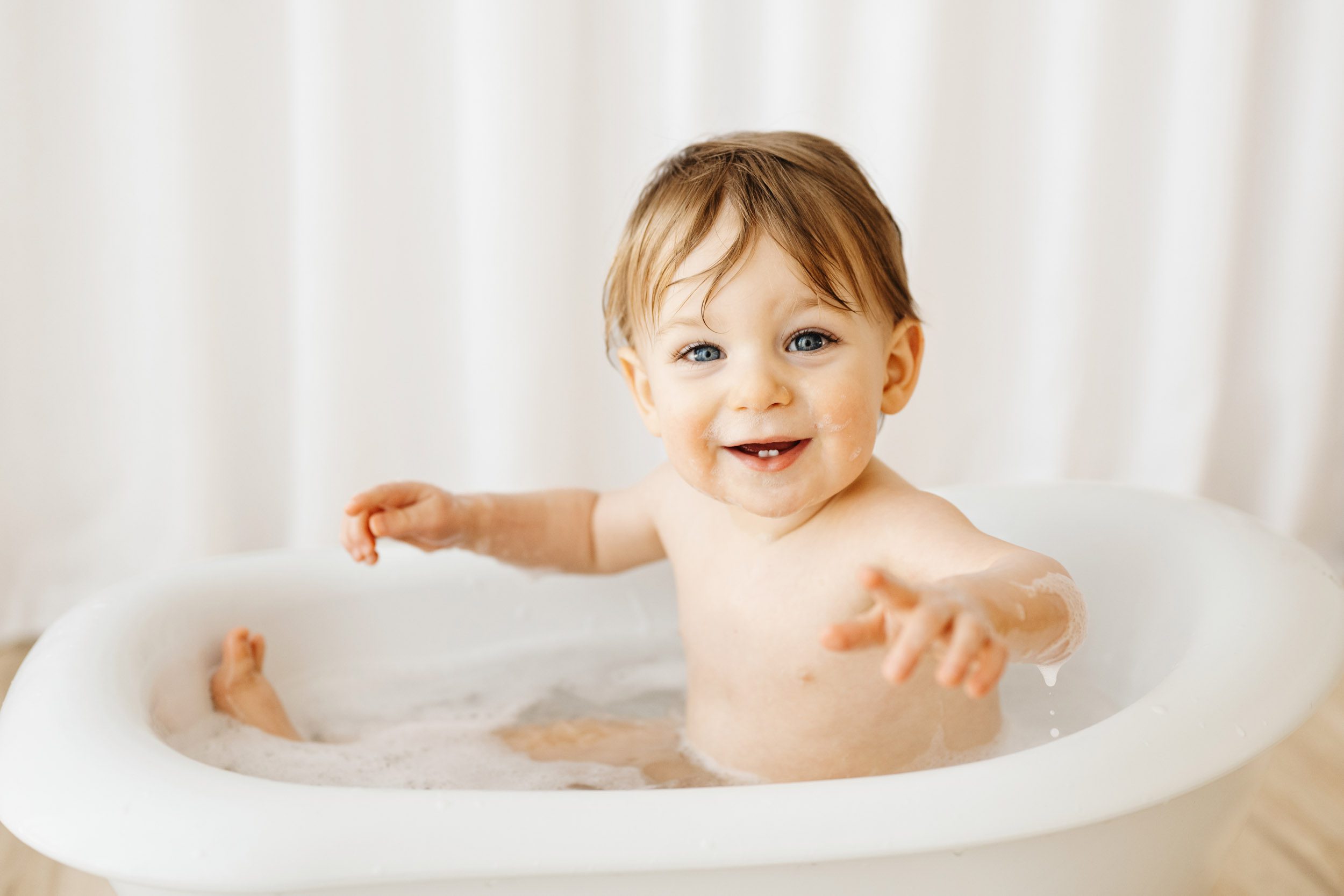 a young boy sitting in a little white bathtub and looking right at the camera with a huge smile on his face during a 1st birthday cake smash & splash photoshoot