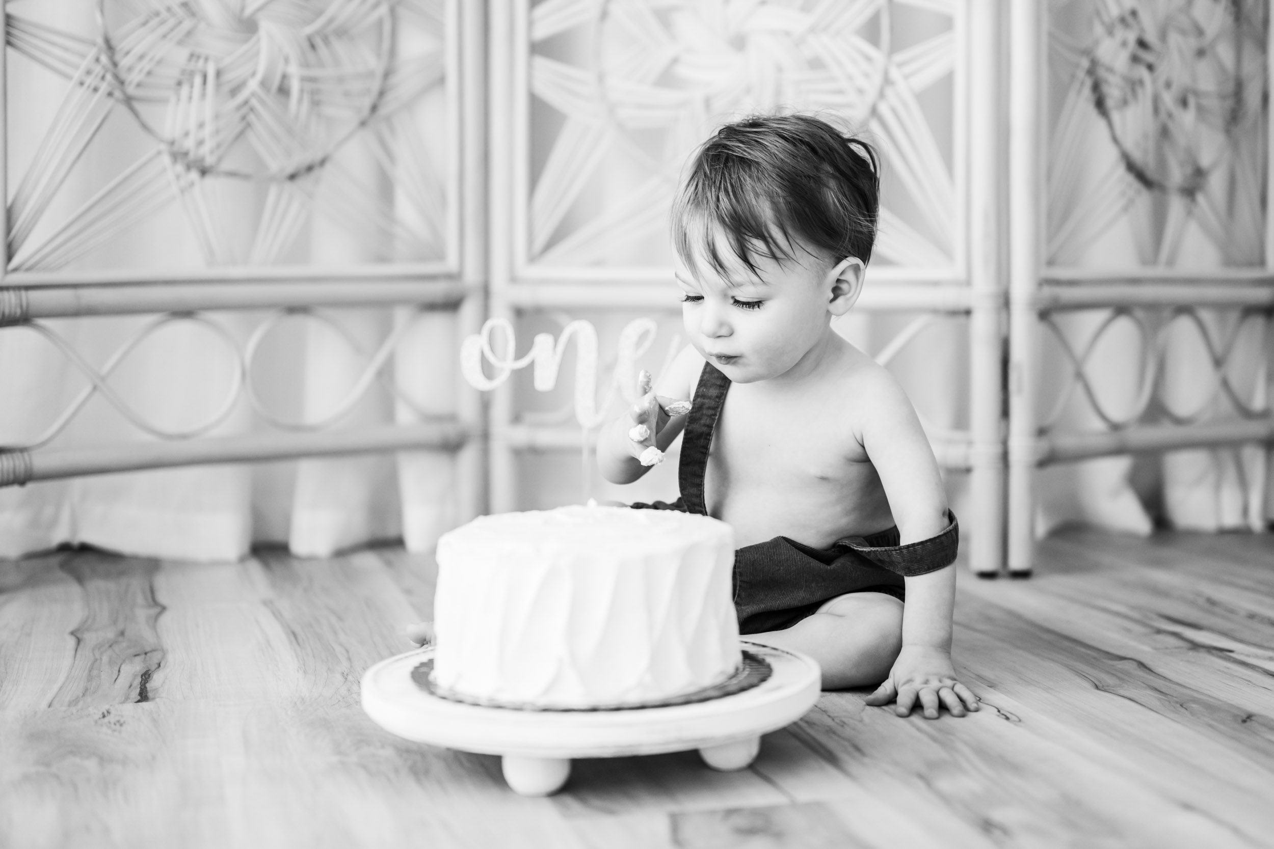 a black and white picture of a young boy sitting on the floor next to a white cake and looking down at his fingers after putting his whole hand in the cake during a 1st birthday cake smash & splash photoshoot
