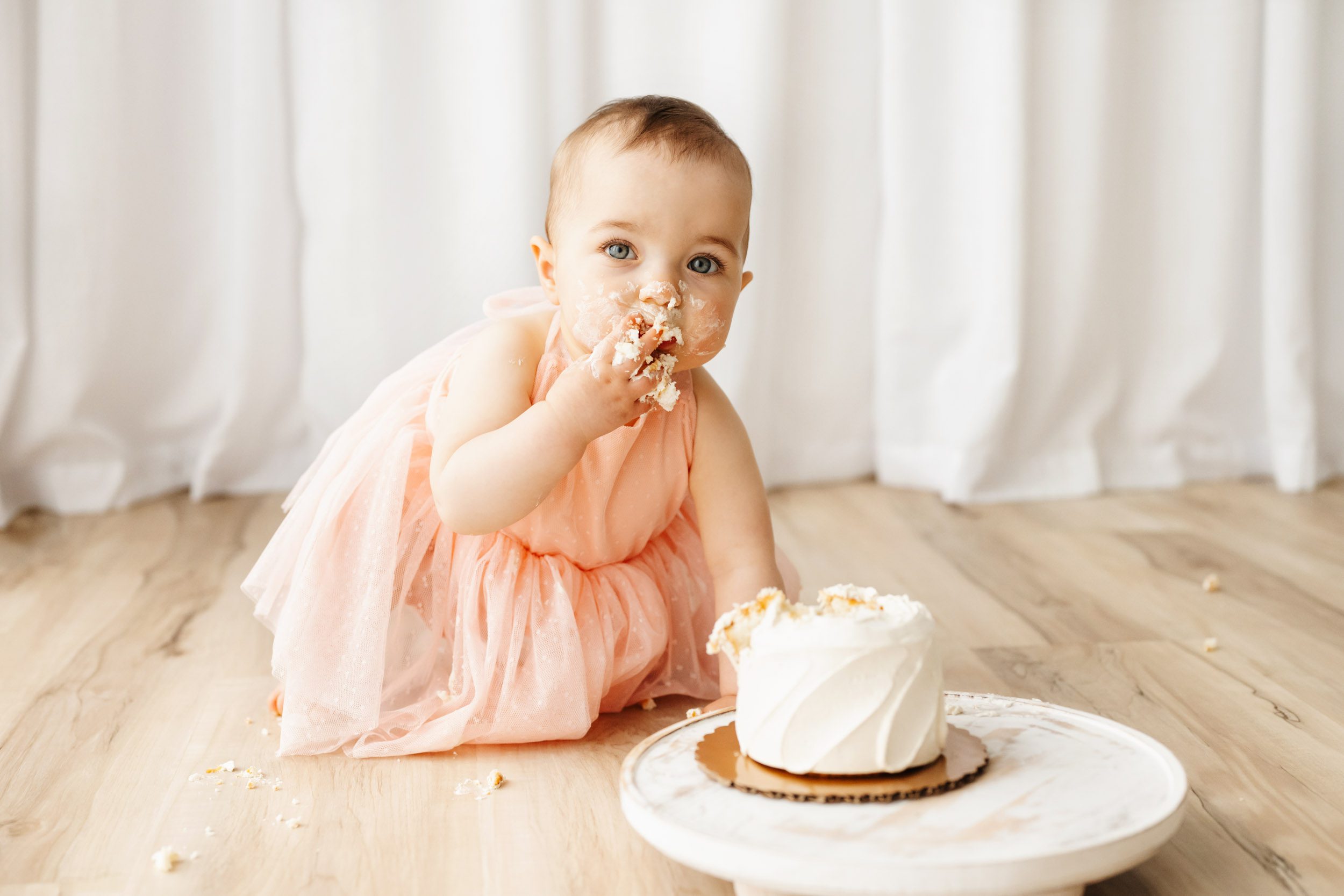 a little girl in a light pink dress crawling across the floor toward a small white cake as eats cake out of her fist during a 1st birthday cake smash photoshoot