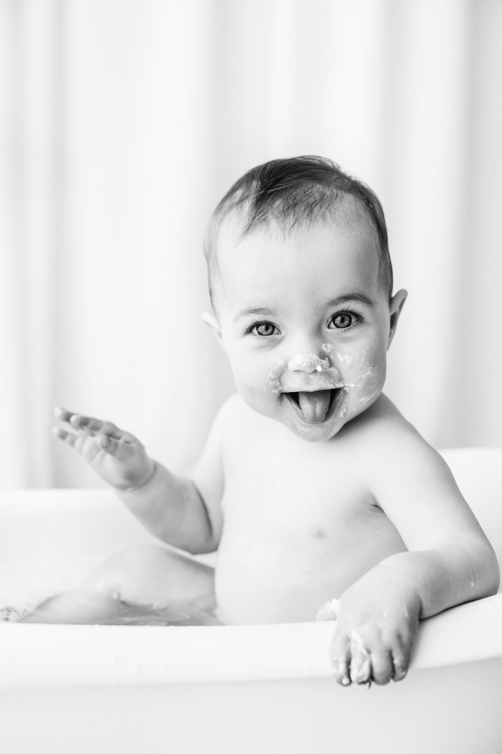 a black and white picture of a little girl with cake all over her face sitting in a little bathtub and sticking her tongue out at the camera during a Pottstown 1st birthday photoshoot 