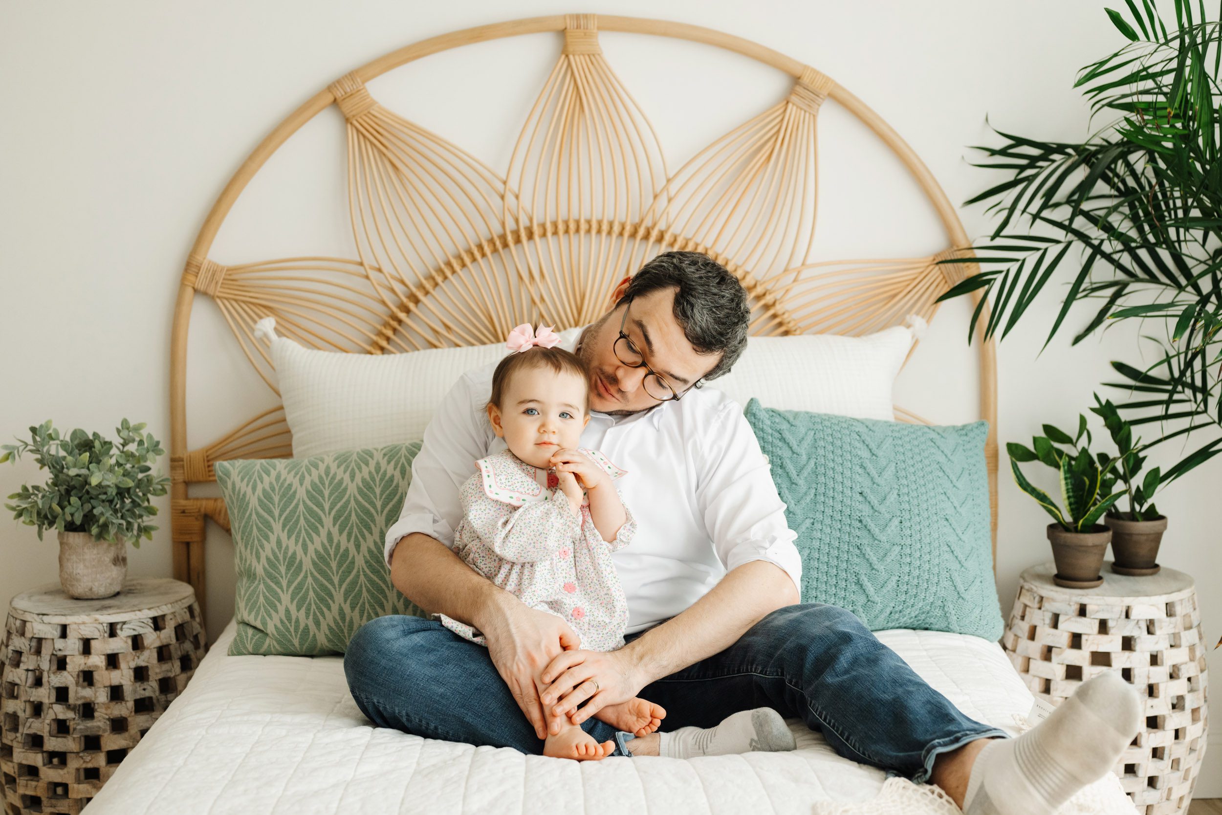 a little girl sitting on her dad's lap on a bed and looking right at the camera while he looks down at her during a 1st birthday photoshoot