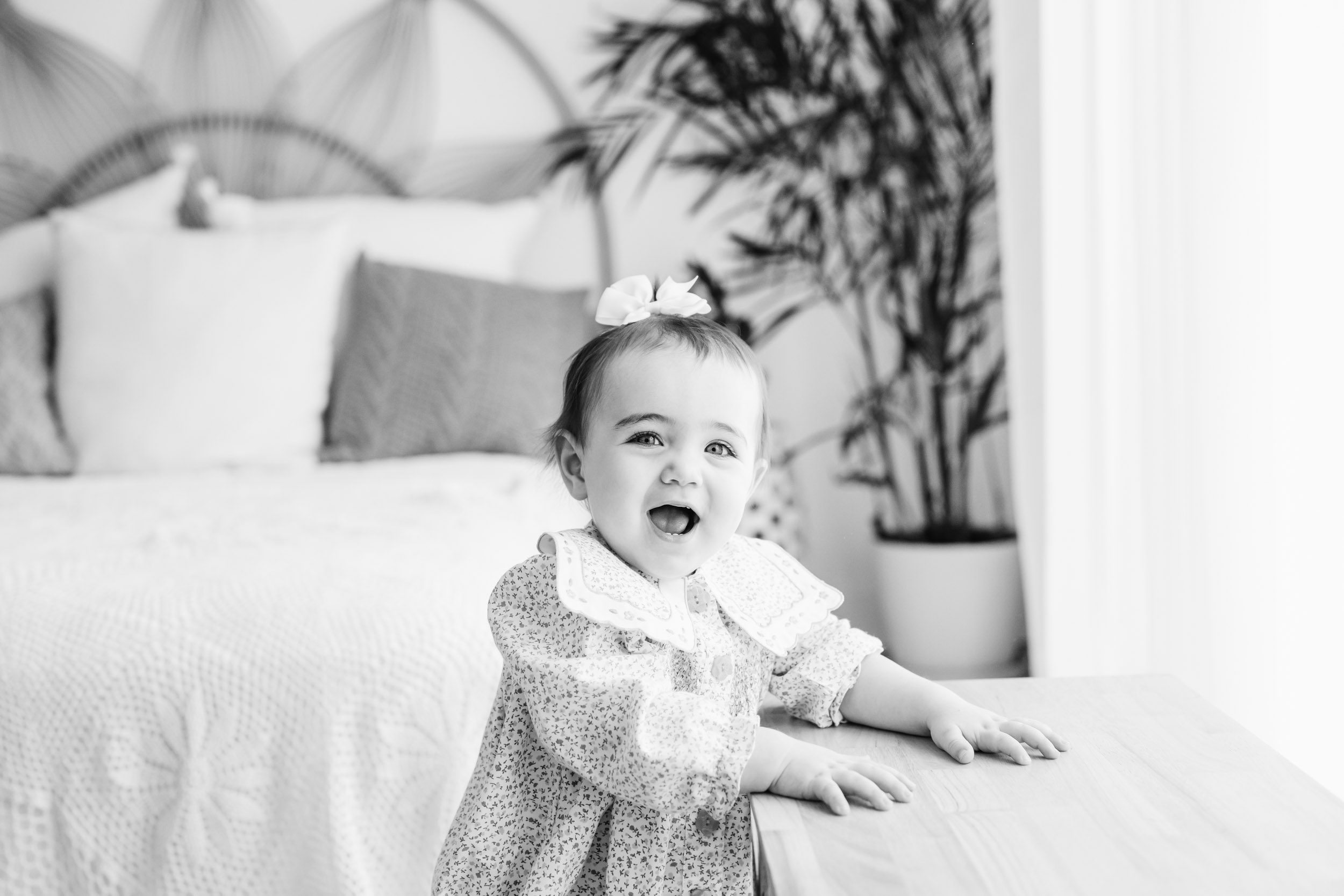 a little girl sitting on a bed and making a surprised face at the camera as she flips through a book laying on the bed in front of her during a 1st birthday photos session