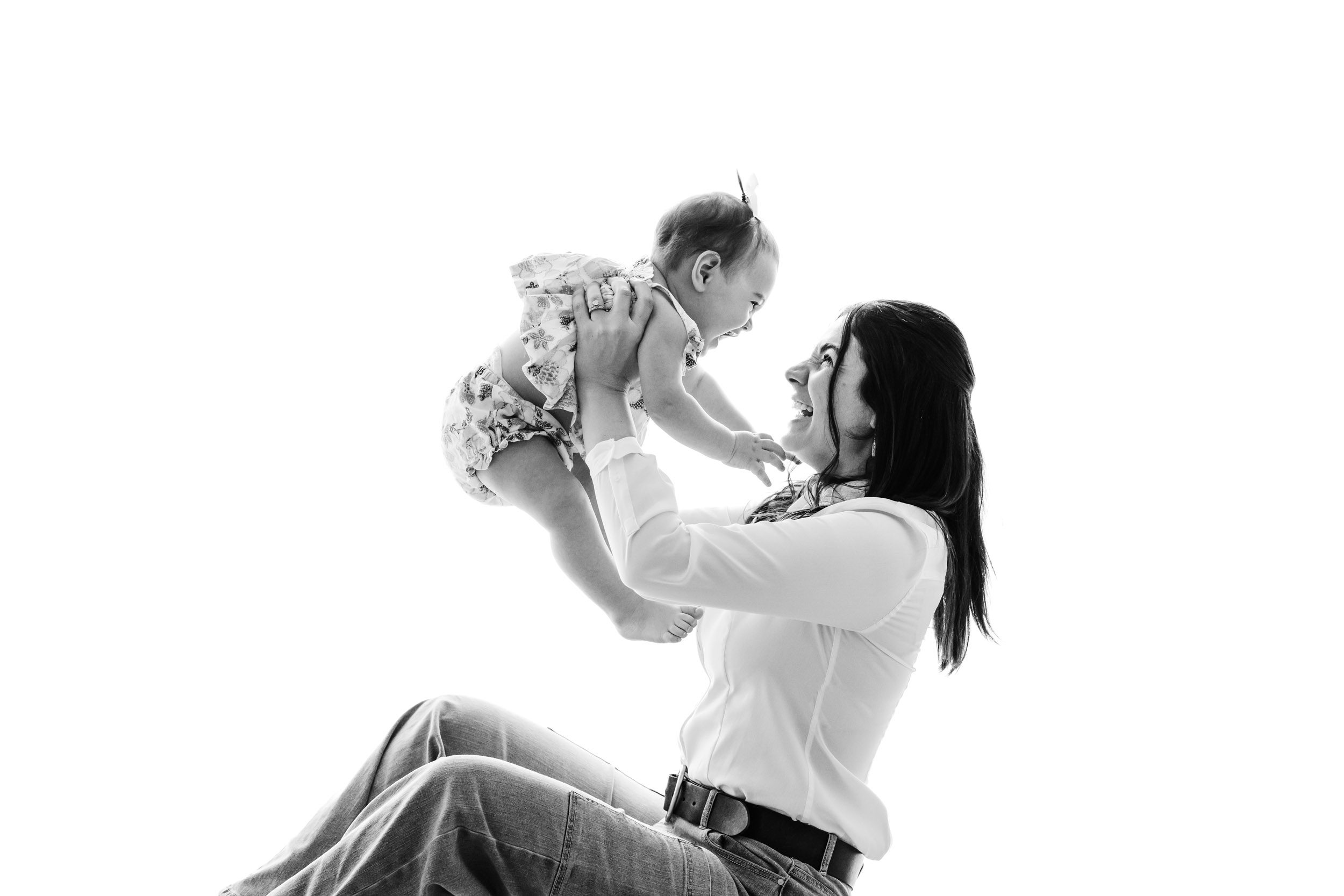 a black and white backlit picture of a mom sitting on a bench in front of a wall of windows and lifting her little girl up in the air as the both laugh during a 1st birthday photos session