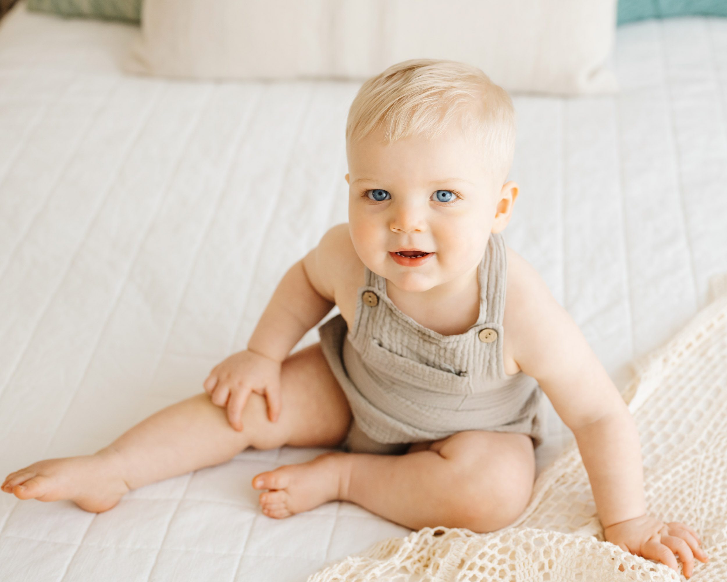 a photo taken from above of a little boy sitting on a bed and looking up at the camera with a shy smile on his face during a first birthday photography session