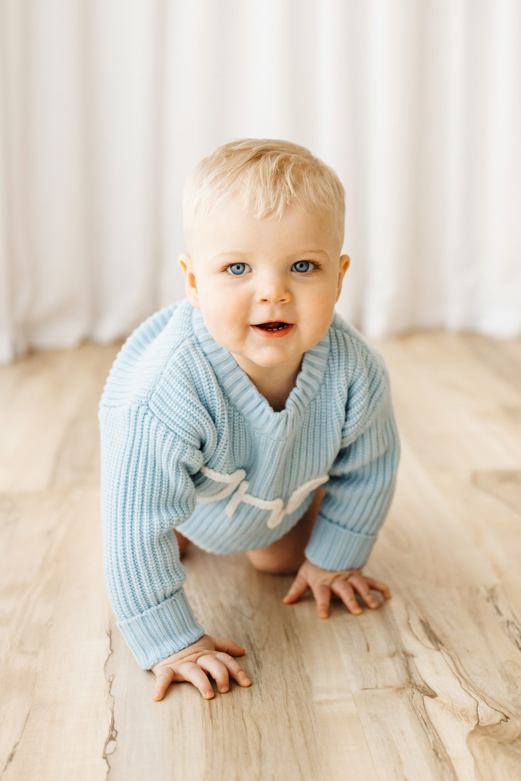 a little boy wearing a light blue knit sweater crawling across the floor as he looks directly at the camera during a first birthday photography session